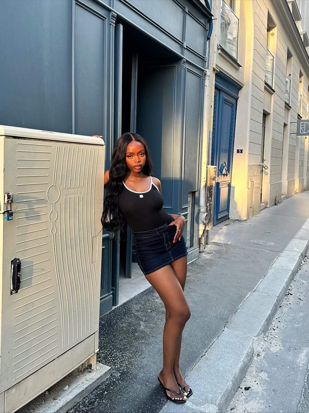 Modelo posa com tamanco de dedo preto em material brilhante, minissaia jeans escura e regata preta com detalhes brancos, em