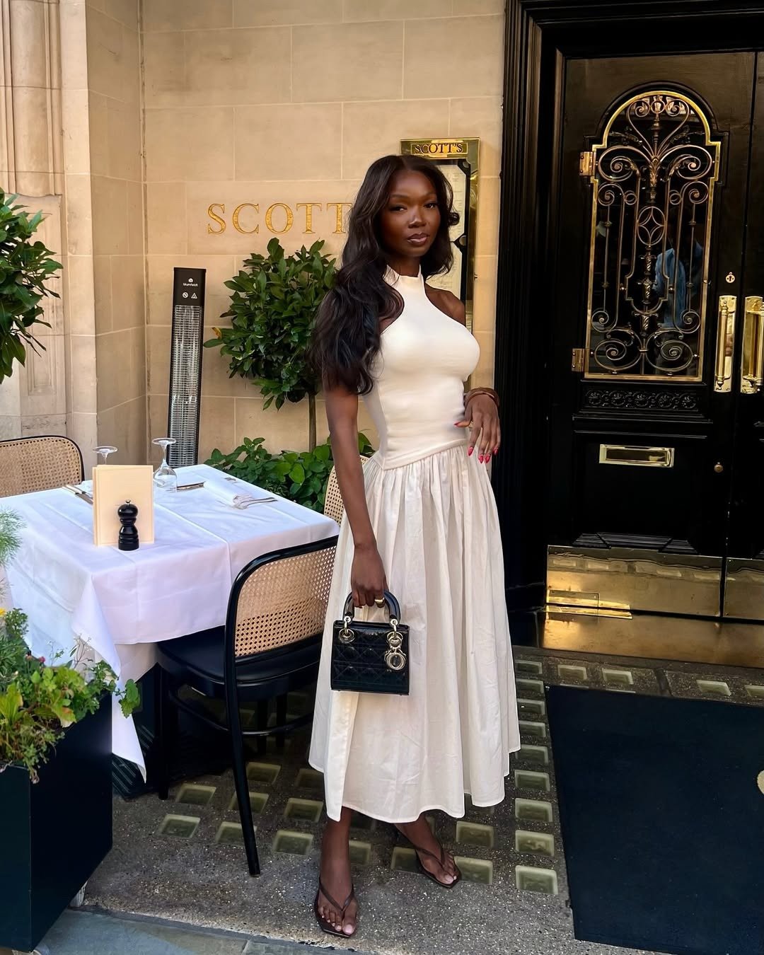 Modelo posa confiante em vestido midi branco com saia rodada e tamanco de dedo marrom, combinando com bolsa preta