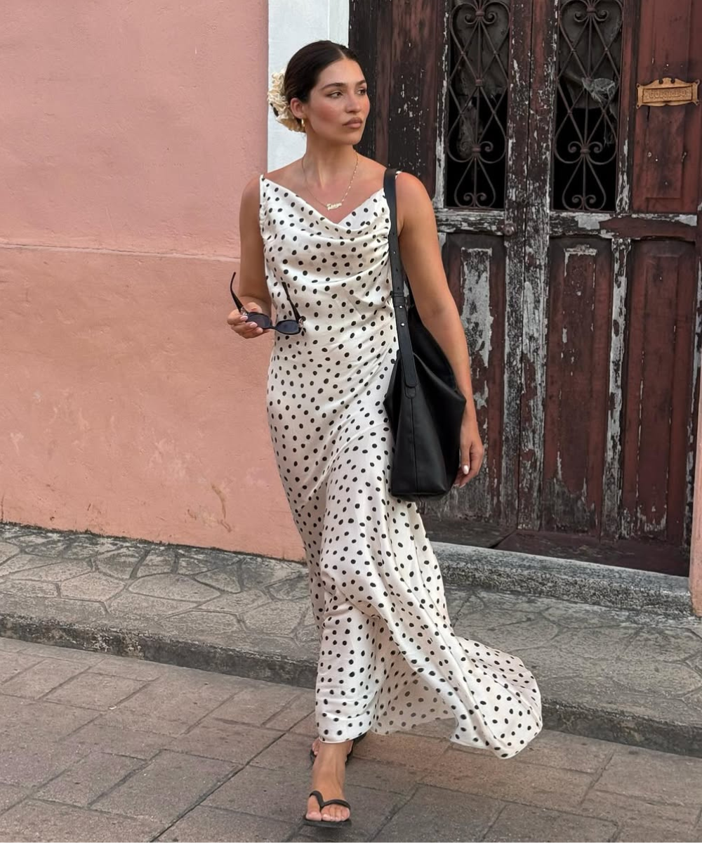 Modelo caminha com vestido longo de tecido fluido branco com poás pretos, combinando bolsa preta e sandálias minimalistas,