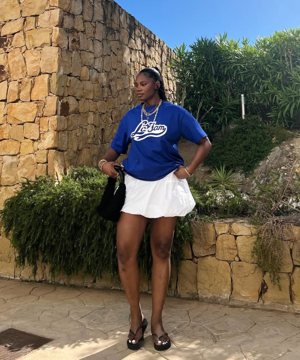 Modelo posa com camiseta azul royal oversized, saia branca bufante e sandálias pretas, transmitindo estilo casual e