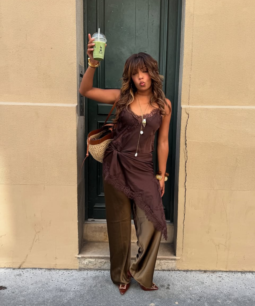 Mulher posa com vestido slip dress marrom de renda sobre calça ampla de cetim e acessórios dourados, transmitindo elegância