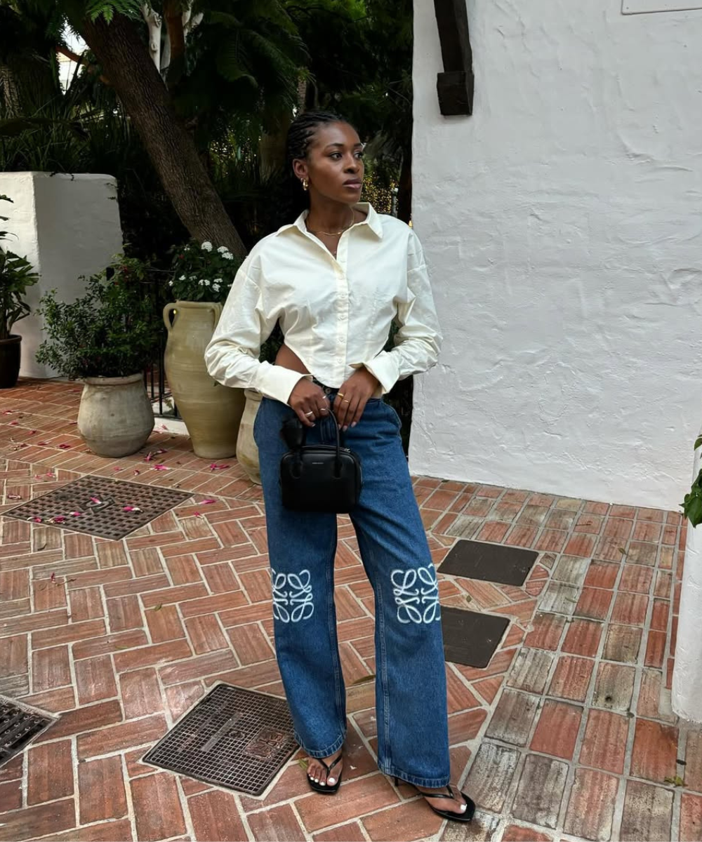 Modelo posa com camisa cropped branca de algodão, jeans azul com bordados brancos e bolsa preta minimalista, transmitindo