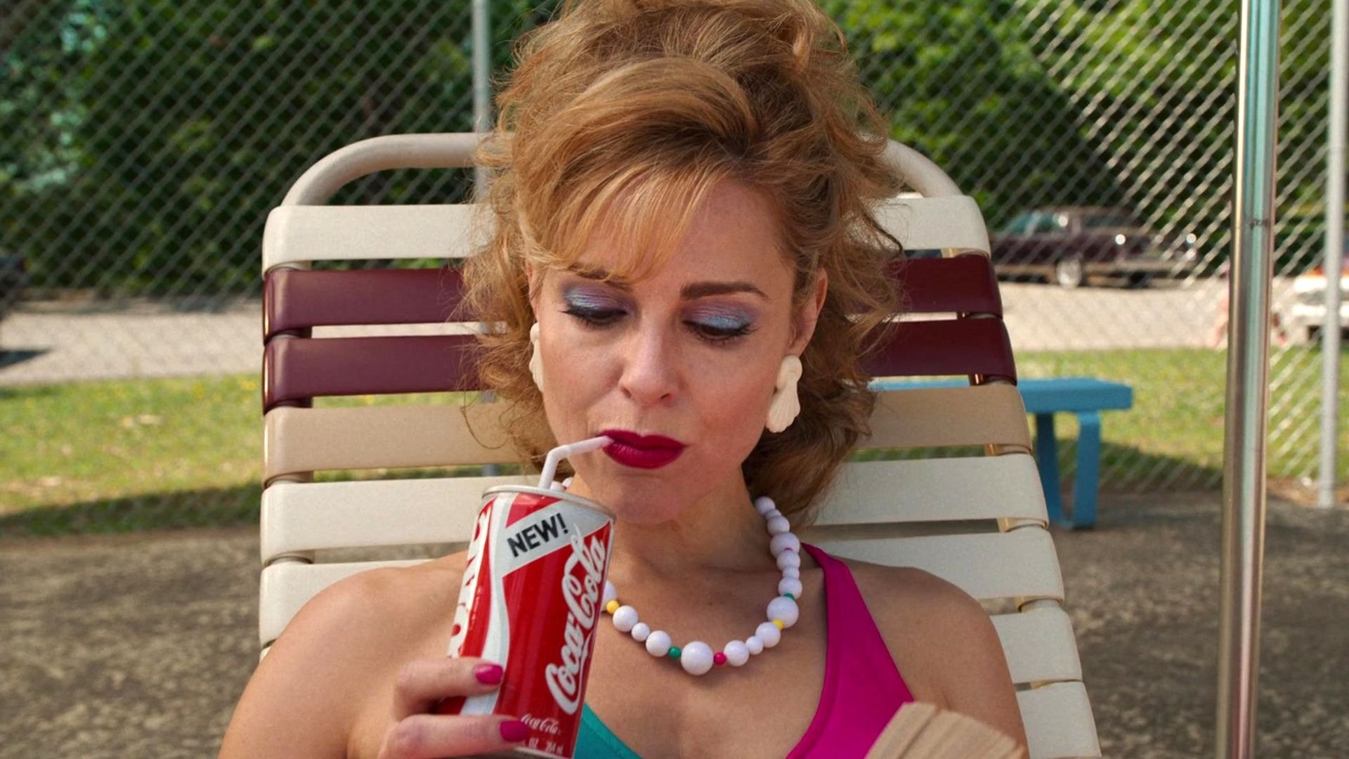 Mulher com maquiagem vibrante e acessórios de resina branca posa relaxada com vestido rosa e colar colorido, refletindo