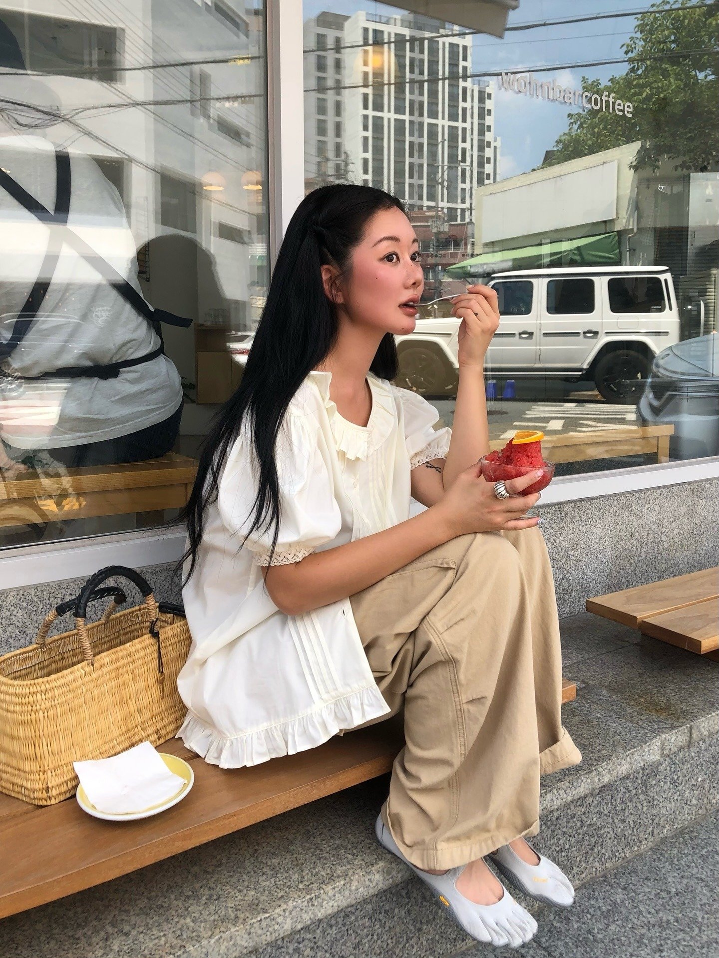 Mulher sentada em um banco, usando uma blusa branca e calças largas, com sapatos de dedos. Ela come um gelato, destacando a tendência dos sapatos feios na moda. Representa tendências de sapatos feios.