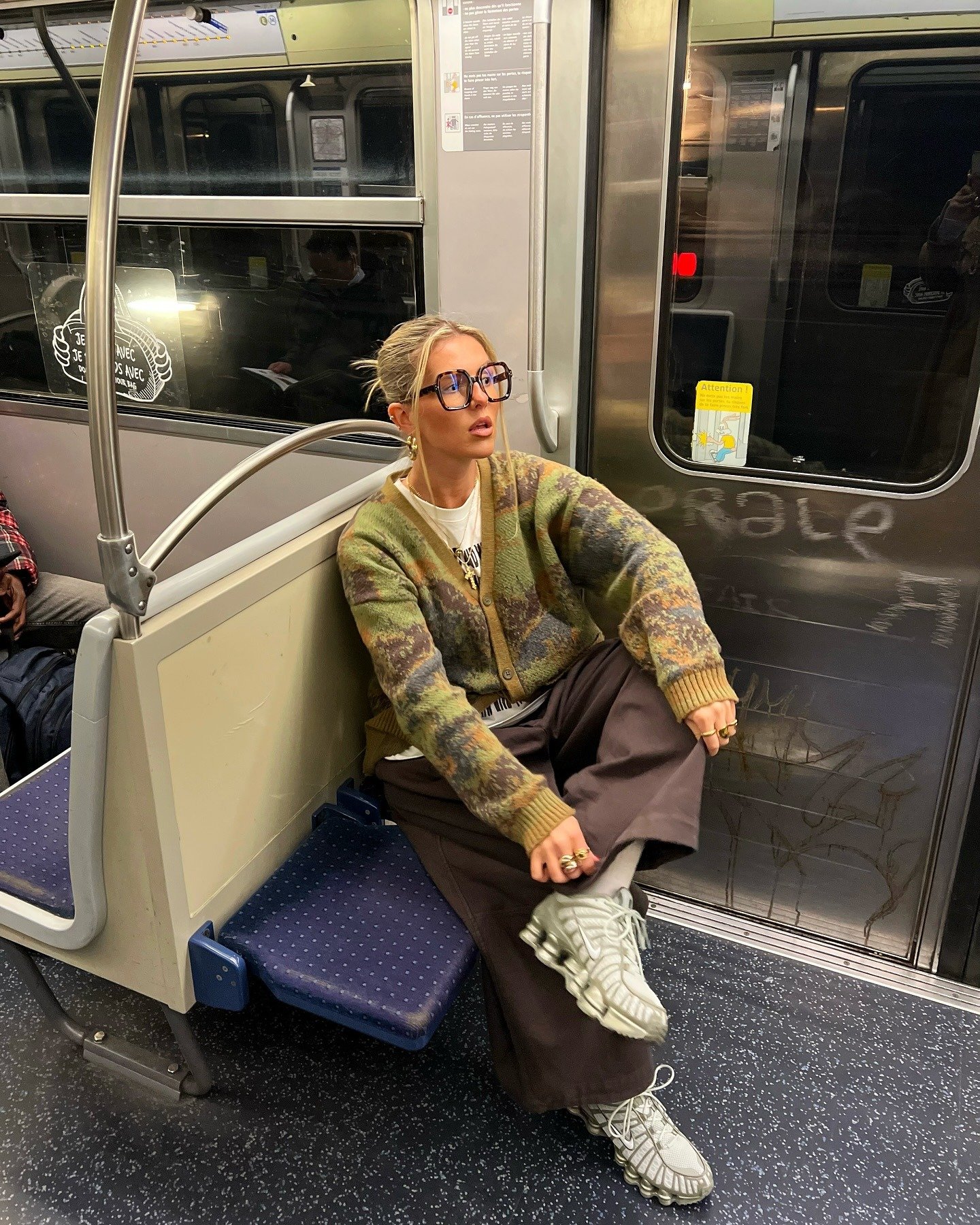A imagem apresenta uma mulher no metrô, usando um suéter oversized e calças largas, destacando sapatos considerados