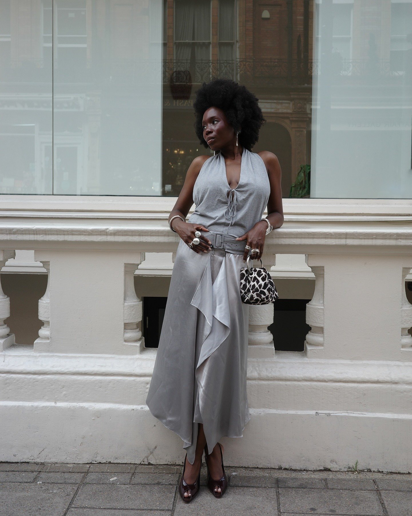 Mulher com cabelo afro usa um vestido cinza fluido, acessórios chamativos e sapatos elegantes, destacando tendências de moda.