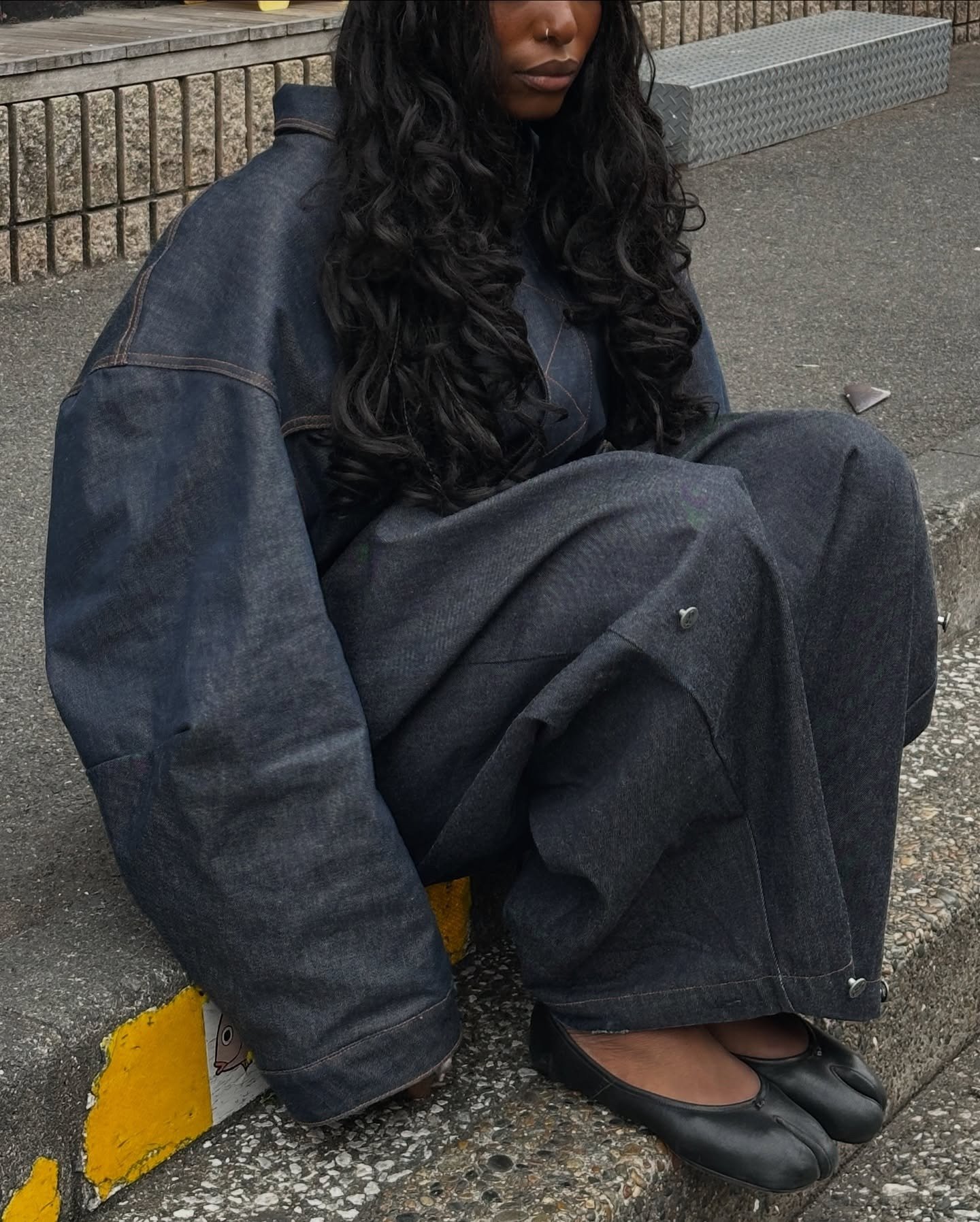 A imagem apresenta uma mulher sentada em uma calçada, vestindo um conjunto oversized de denim, que reflete a tendência de roupas largas e confortáveis. Seus sapatos pretos, que podem ser considerados