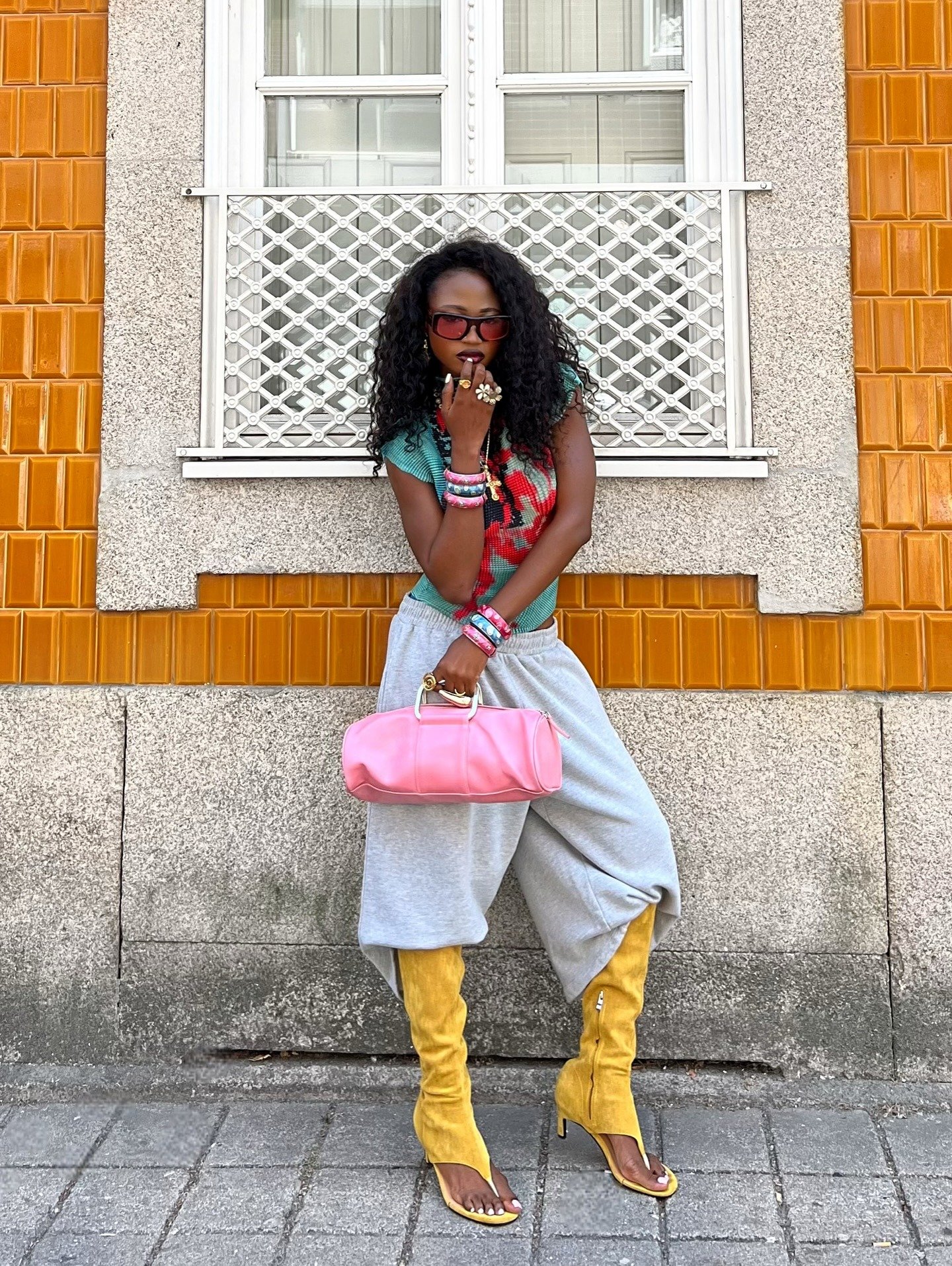 Mulher com cabelo cacheado, usando blusa colorida, calça cinza larga e botas amarelas de salto alto. Acessórios incluem pulseiras e bolsa rosa. O look combina elementos ousados, refletindo as tendências de sapatos feios, destacando a originalidade na