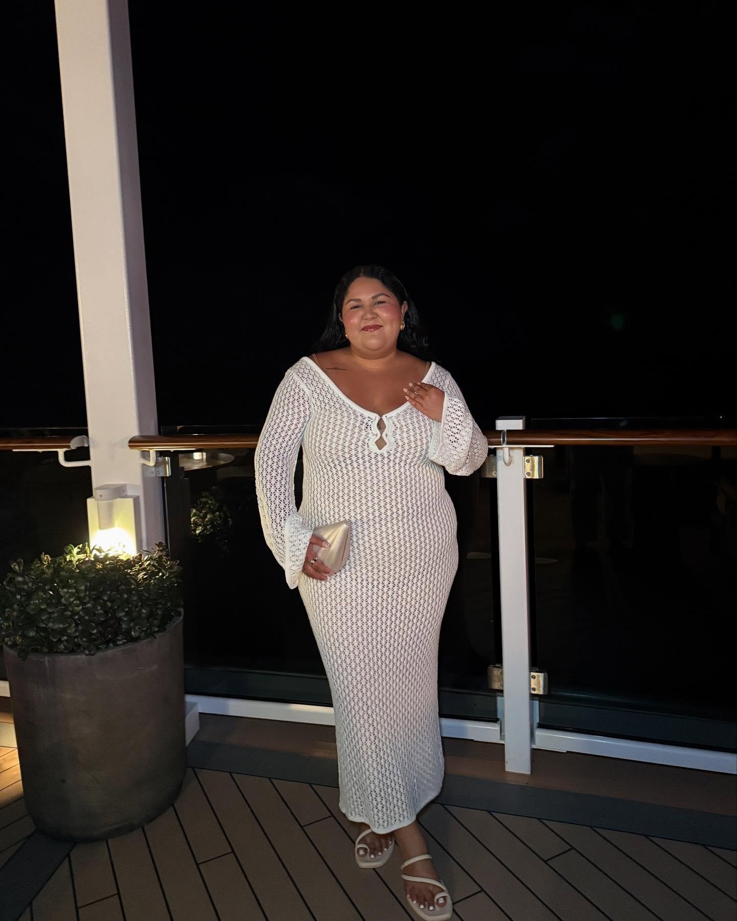 Modelo posa sorridente com vestido branco de tricô ajustado, mangas amplas e decote com recortes, clutch perolada e