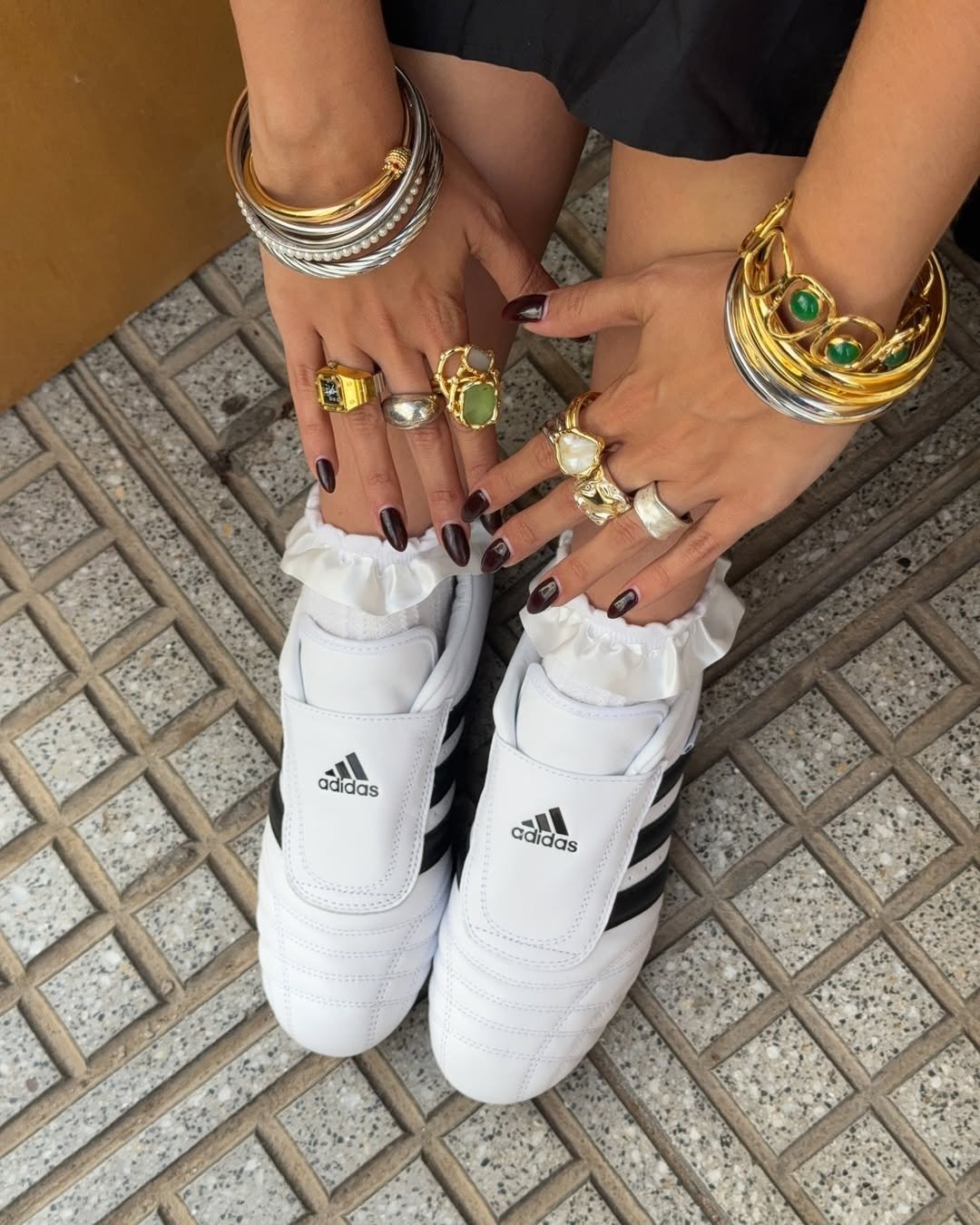 Tênis slip on branco Adidas com listras pretas, combinado com meias de babado e joias douradas e prateadas, estilo moderno e