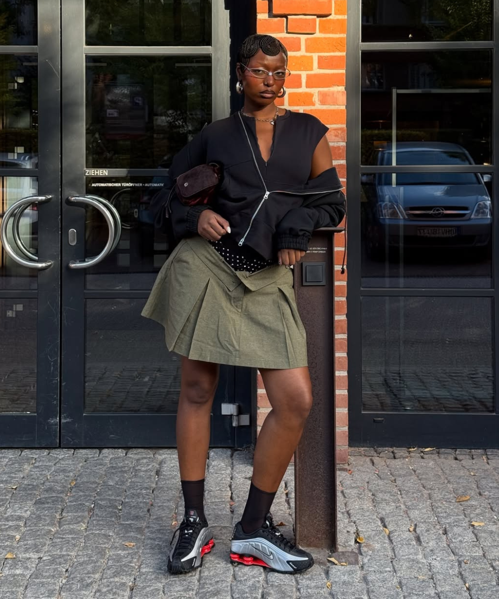 Modelo posa com tênis tendência de 2026 em preto, cinza e solado vermelho, combinando saia plissada verde e jaqueta preta