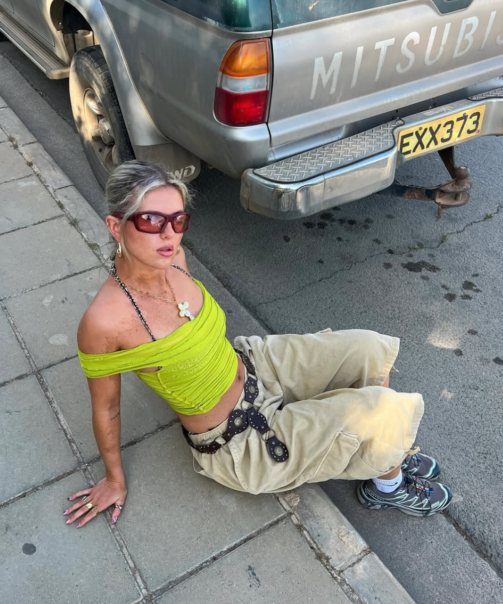 Modelo senta no chão urbano com top verde limão ajustado, calça cargo bege ampla, cinto de fivela larga e tênis tendência de