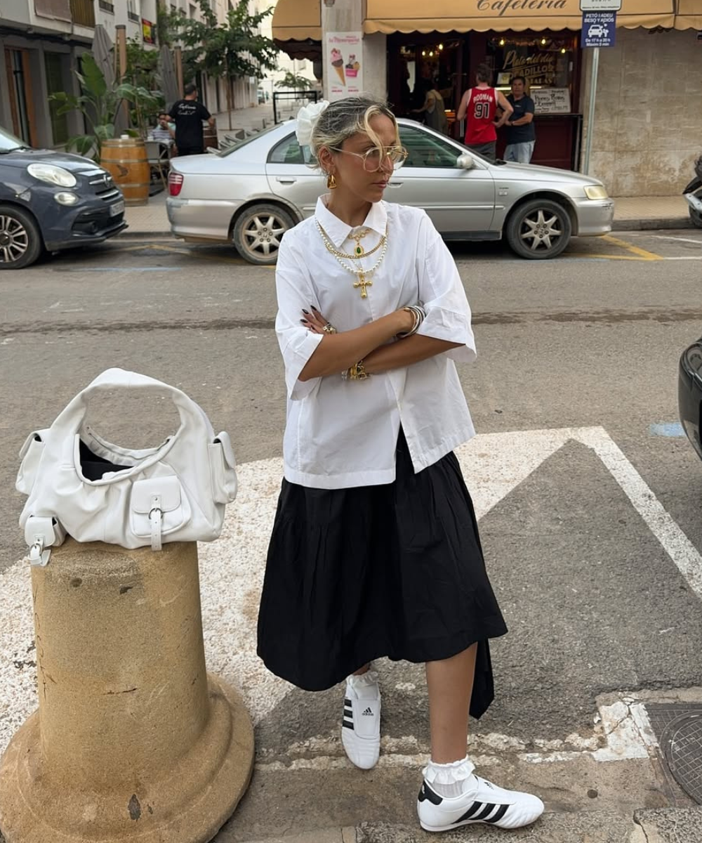 Modelo posa com camisa branca oversized, saia preta fluida e tênis tendência de 2026 branco com listras pretas, estilosa.