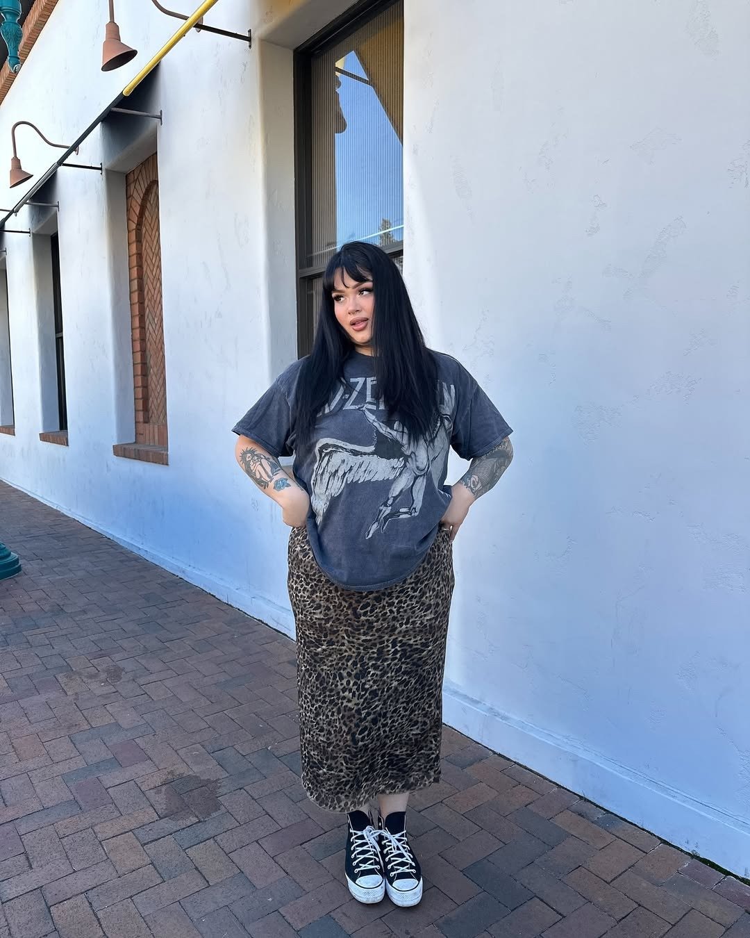A imagem apresenta uma jovem com estilo descontraído, usando uma camiseta oversized de banda e uma saia longa com estampa de onça. Nos pés, um par de tênis pretos, ideais para o verão, combinando conforto e moda. Tênis para o verão