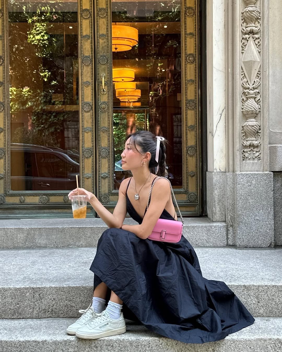 A imagem retrata uma jovem sentada em uma escada, usando um vestido preto longo e elegante, ideal para o verão. Seus tênis brancos, combinados com meias brancas, trazem um toque casual ao look. Tênis para o verão.