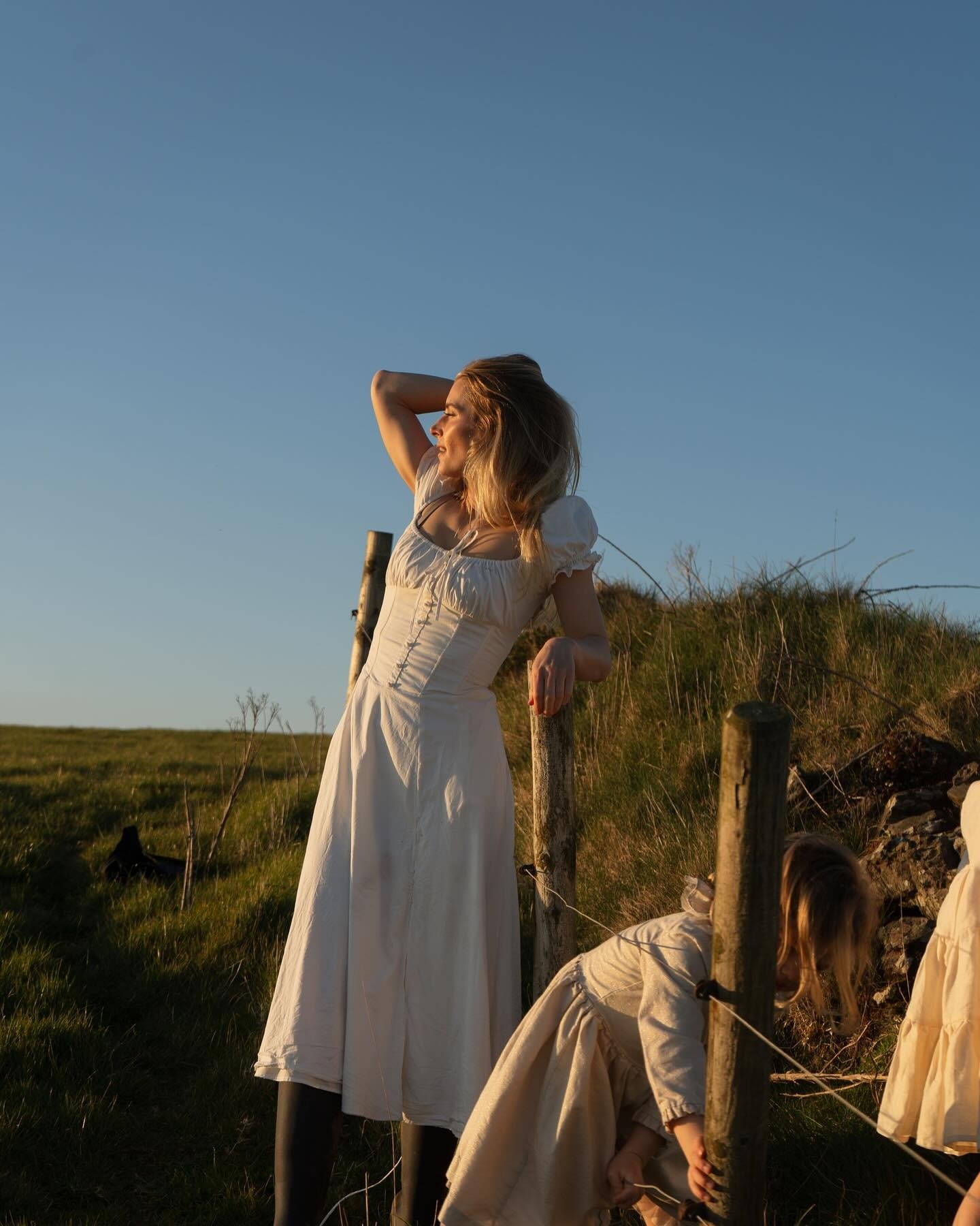 Mulher em vestido branco de algodão com mangas bufantes e saia midi, posa relaxada ao pôr do sol em campo, estilo tradwife.