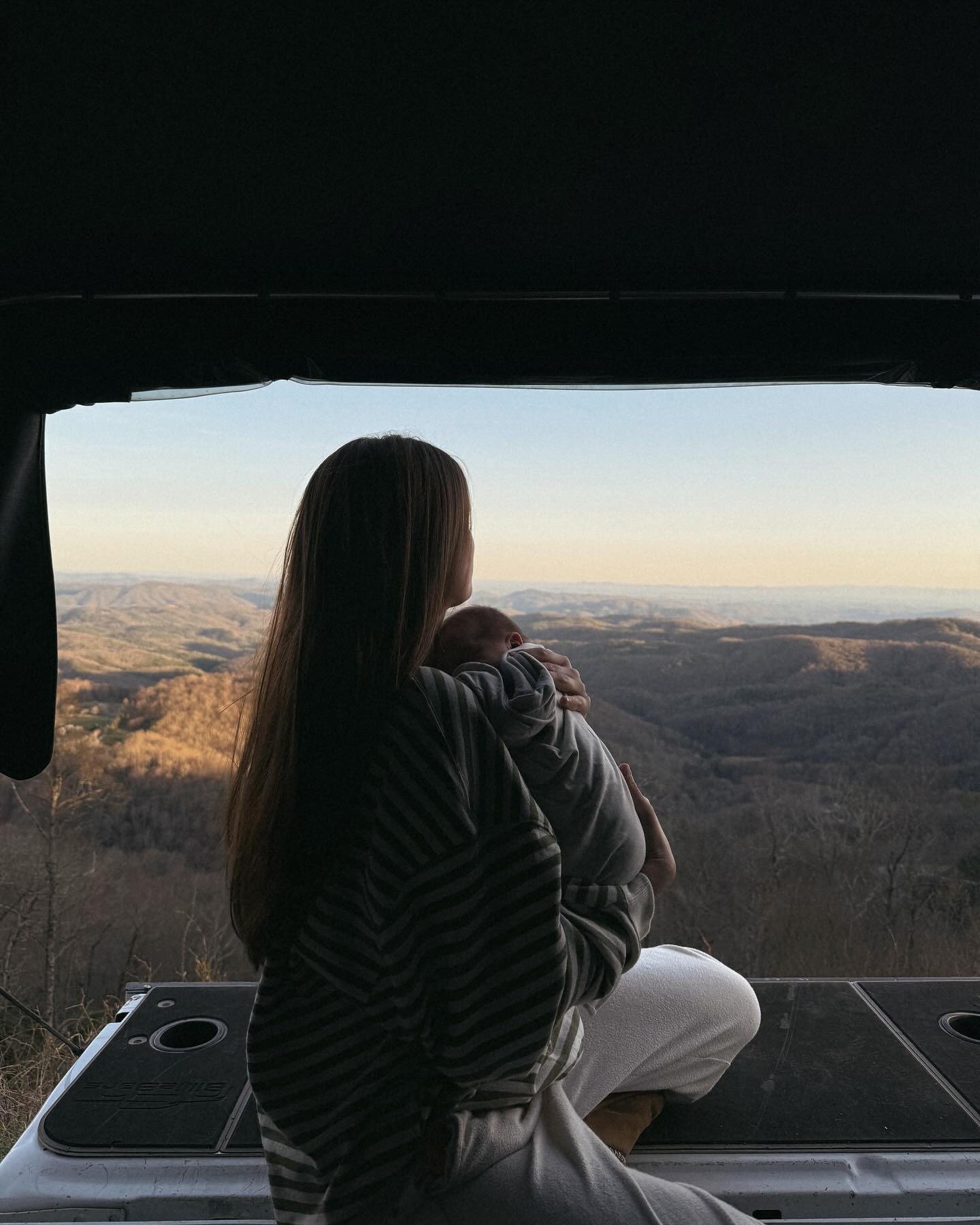 Mulher tradwife segura bebê com roupa confortável em tons neutros, sentada com vista para montanhas ao entardecer.
