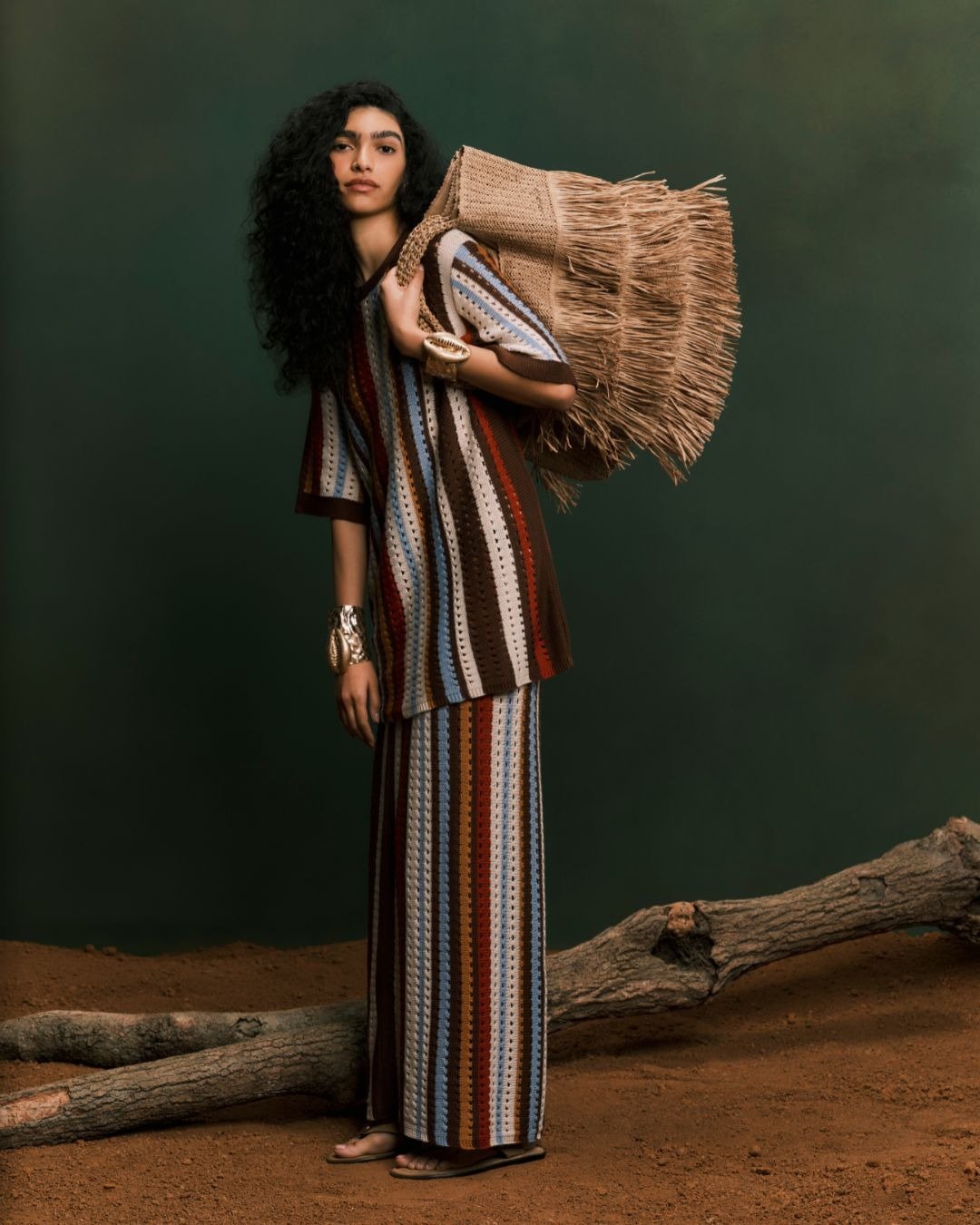 Modelo em tricô de verão, vestido listrado em tons terrosos e azuis, com acessórios em metal e bolsa de palha. O fundo verde destaca a leveza e frescor da moda de verão, ideal para looks casuais e sofisticados.