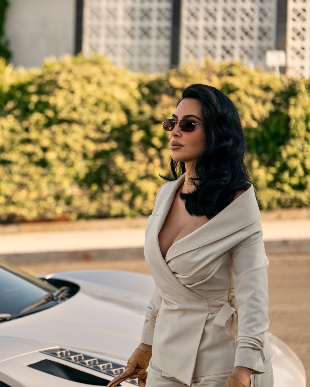 A série da Kim Kardashian destaca momentos de elegância e estilo. Na imagem, ela exibe um look sofisticado com um traje bege, que valoriza suas curvas, e óculos escuros modernos. O cenário, com vegetação ao fundo, complementa a estética.