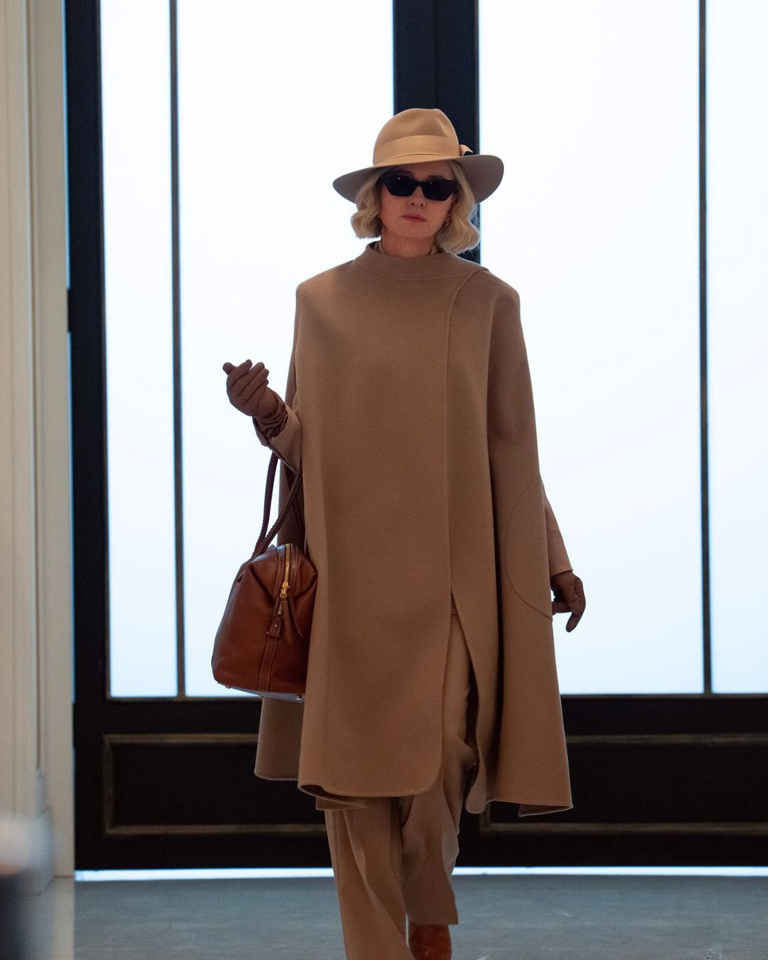 Na série da Kim Kardashian, uma mulher elegante caminha com confiança, vestindo um conjunto monocromático em tons de bege. O look inclui um casaco de linhas minimalistas, calças largas e um chapéu estiloso.