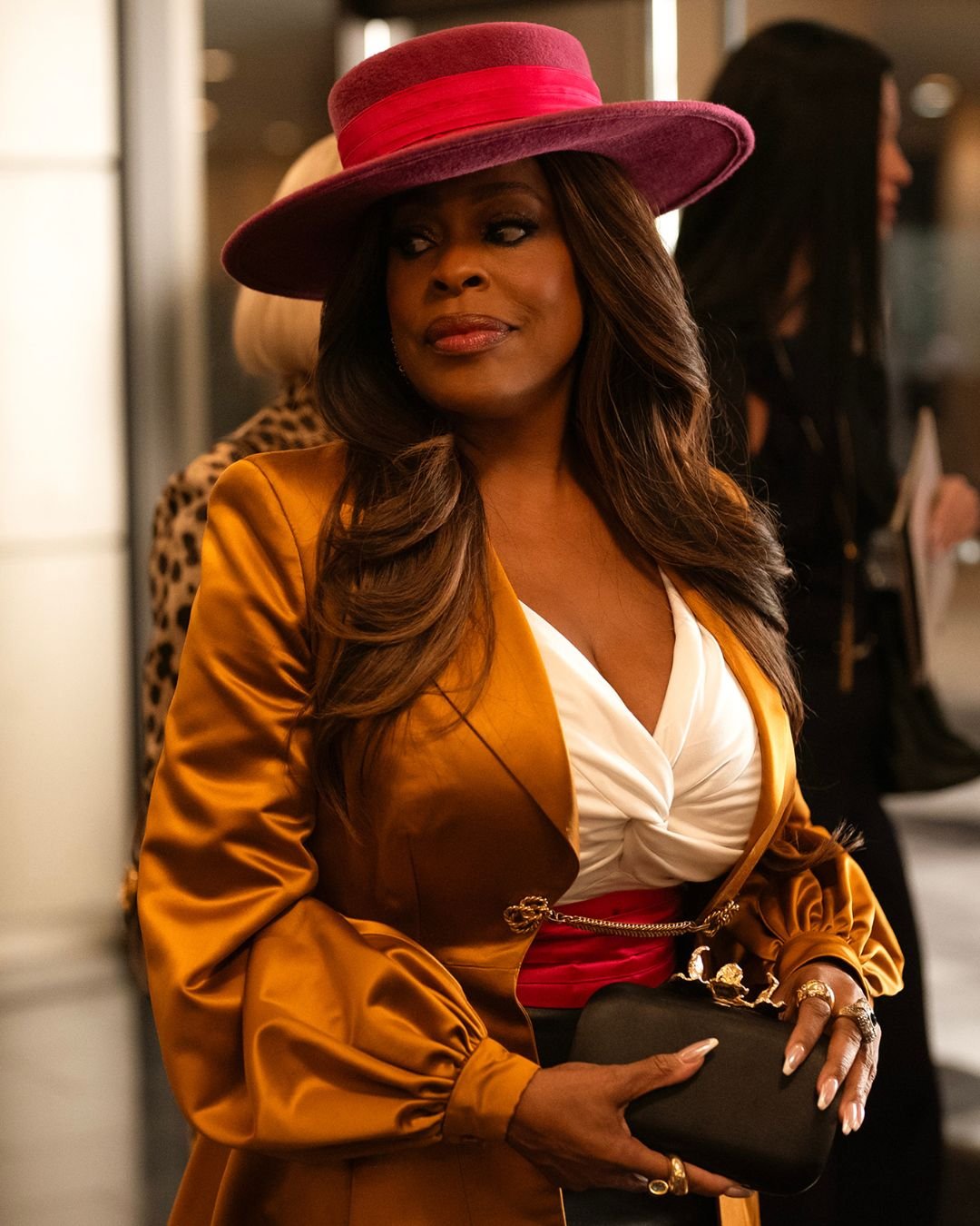 Personagem de Tudo é Justo, série da Kim Kardashian, usando um blazer dourado com mangas volumosas, destacando um cinto vermelho que acentua a cintura. Ela complementa o look com uma blusa branca e um chapéu rosa vibrante.