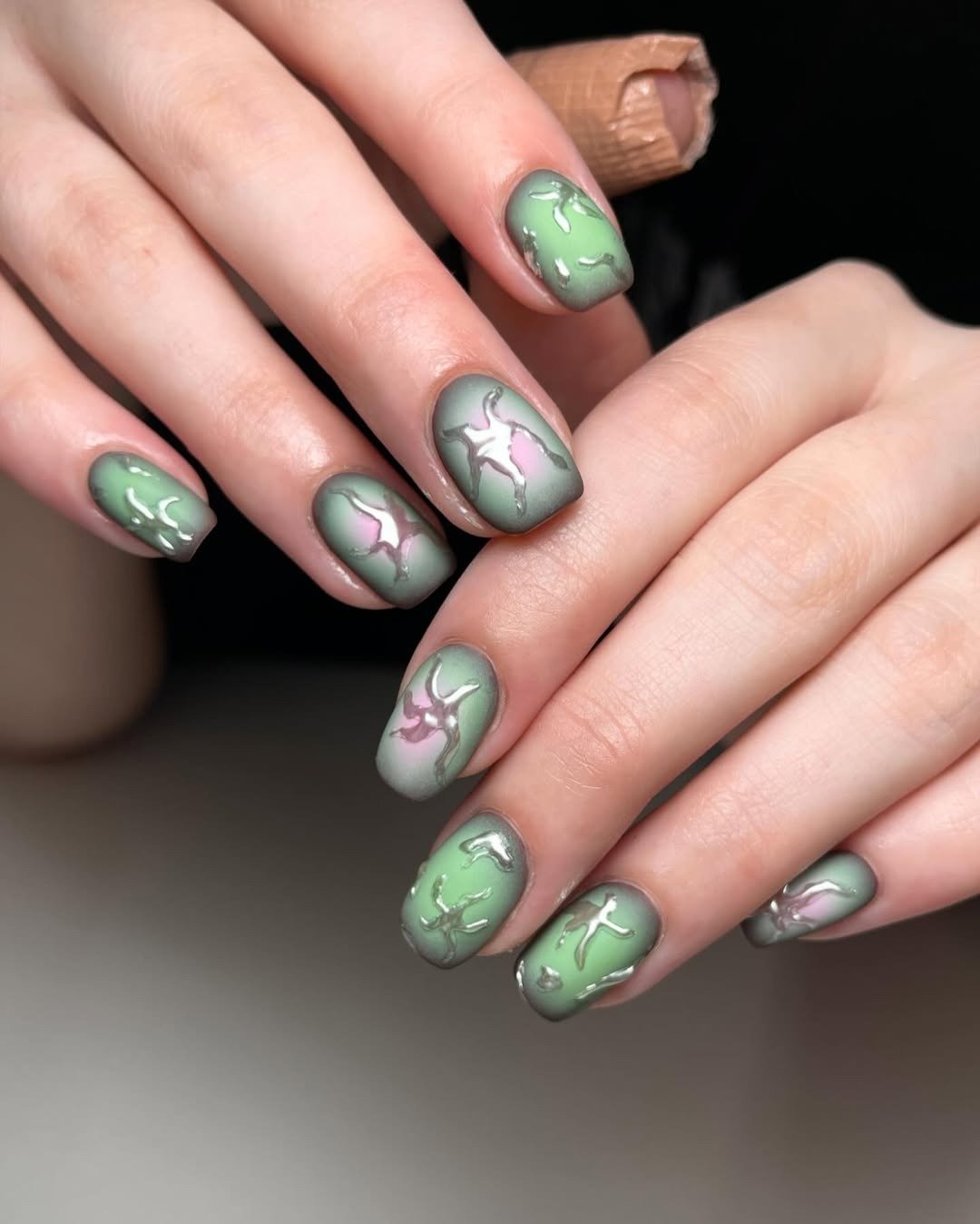 Unhas com esmalte fosco em tons de verde e lilás, com detalhes em relevo prateado que criam efeito artístico e moderno.