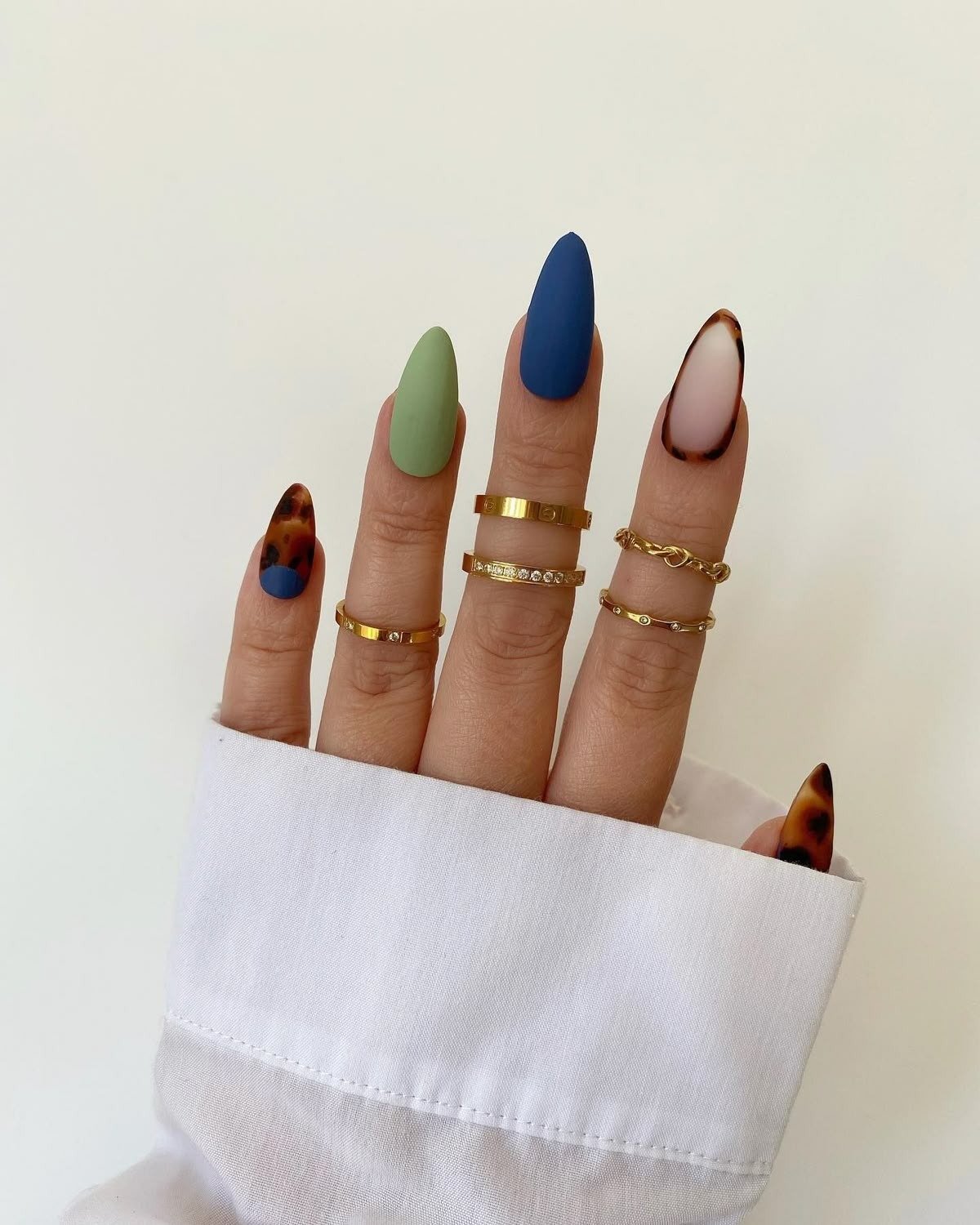 Unhas longas com esmalte fosco verde e azul, detalhes em animal print e anéis dourados delicados criam estilo moderno.