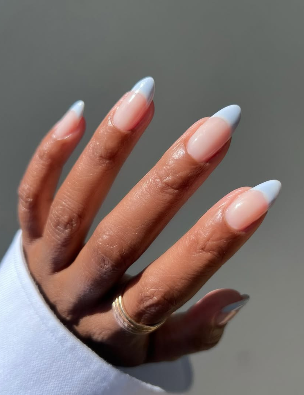Unhas para o Ano Novo apresentam um design elegante com formato stiletto e uma combinação de tons nude e azul claro nas pontas. A pele das mãos está bem cuidada, destacando o brilho das unhas. Um anel delicado complementa o visual.