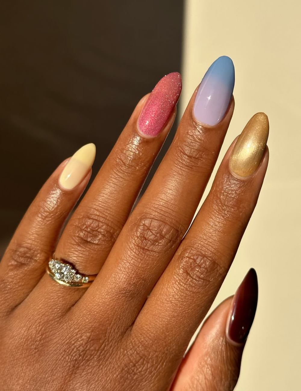 As unhas para o Ano Novo apresentam um design moderno e vibrante. Cada unha exibe uma cor diferente: um tom nude suave, um rosa brilhante com glitter, um azul degradê, um dourado metálico e um vermelho profundo.