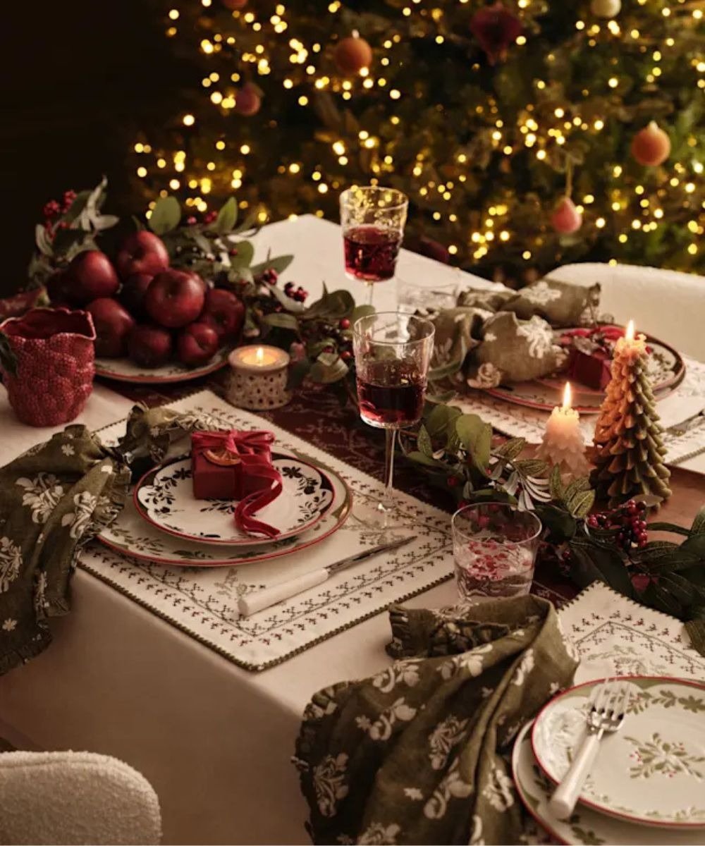 Mesa de natal simples e bonita com toalha branca, louças florais, guardanapos verdes, velas temáticas e maçãs vermelhas.