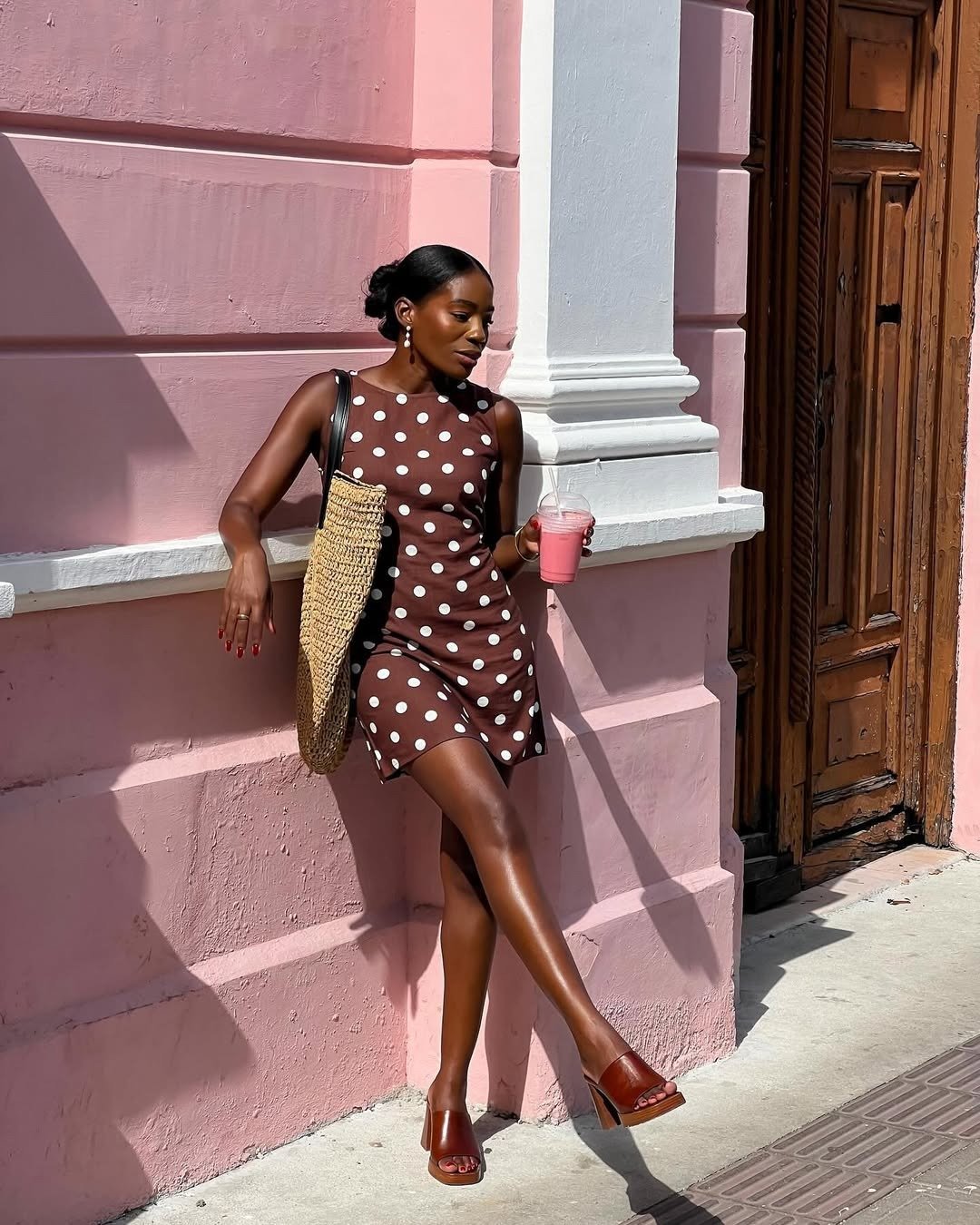 Modelo posa com vestido feminino curto marrom de poás brancos, tecido leve, sandálias caramelo e bolsa de palha, em clima