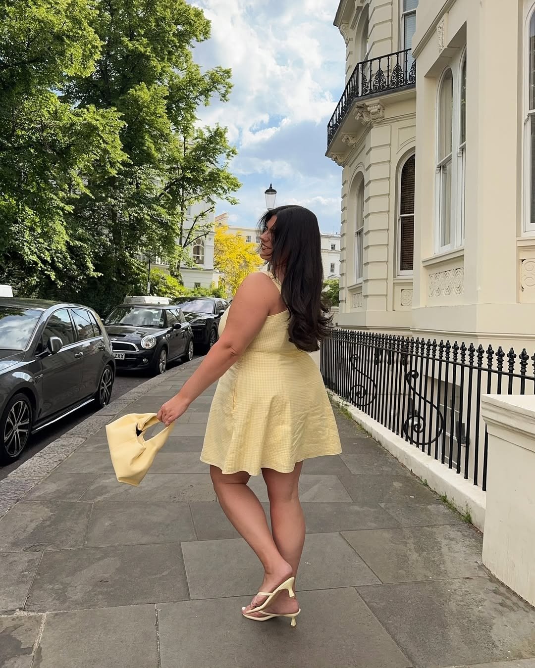 Vestido feminino curto amarelo com modelagem evasê em tecido leve, modelo posa sorrindo ao ar livre com bolsa e sandália.