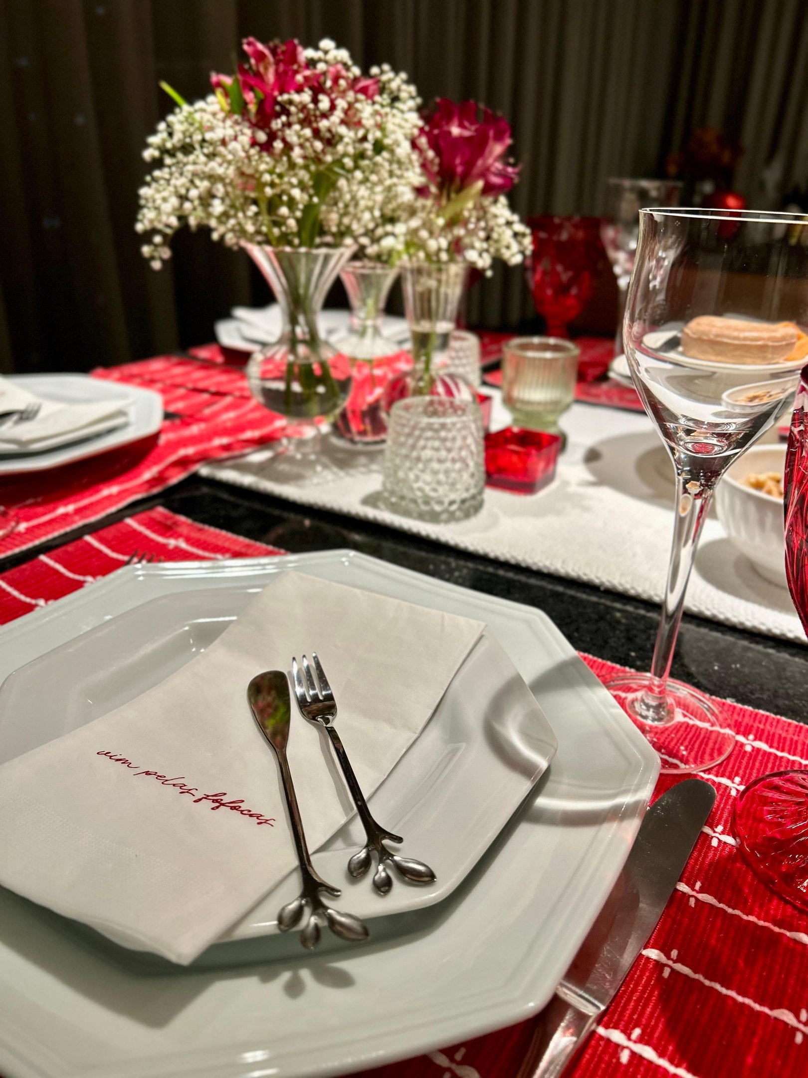 Mesa posta para Natal em casa com pratos brancos octogonais, talheres delicados, guardanapo bordado e detalhes em vermelho.