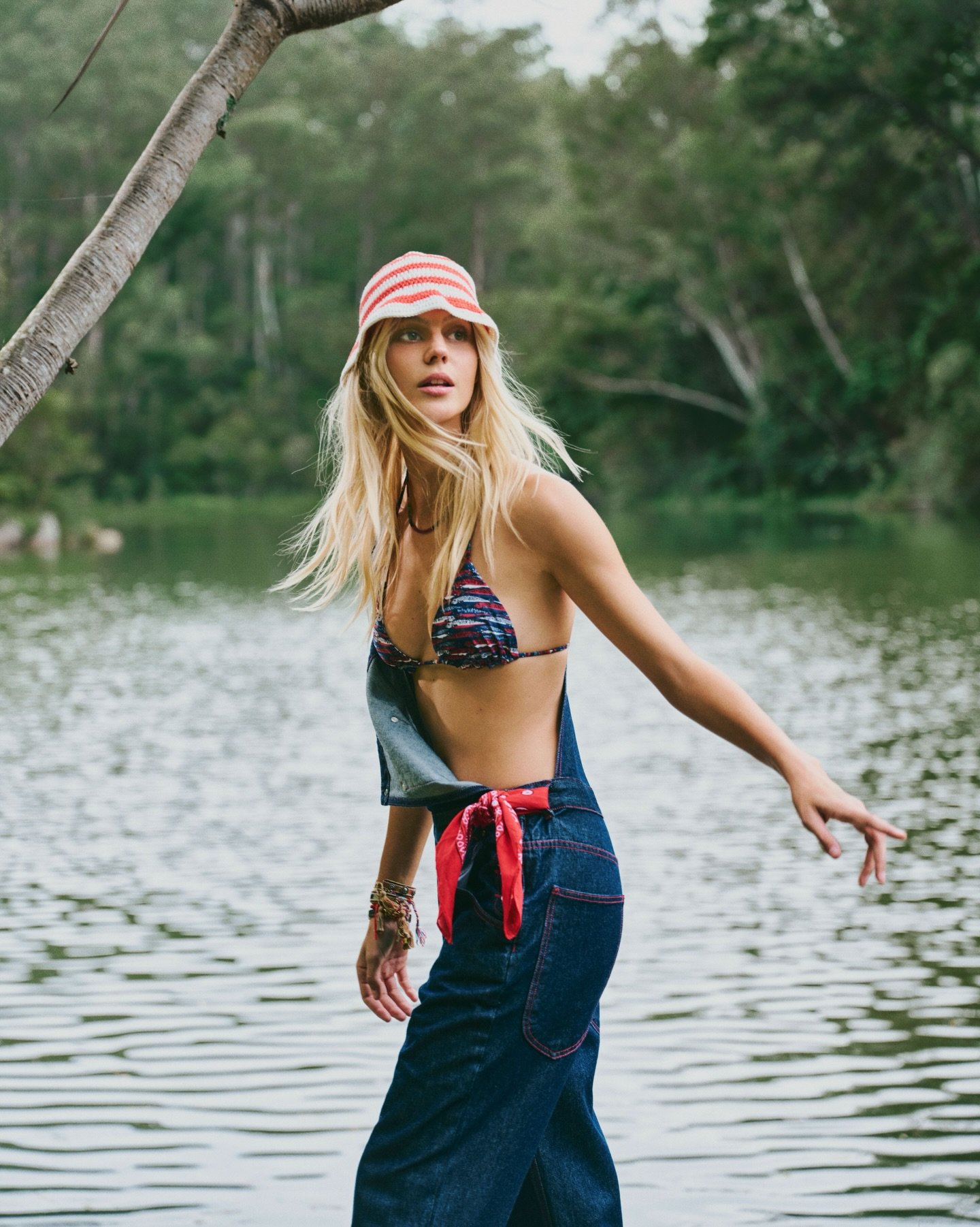 Modelo posa junto a lago vestindo top de biquíni estampado, jardineira jeans com lenço vermelho e chapéu listrado, estilo NV