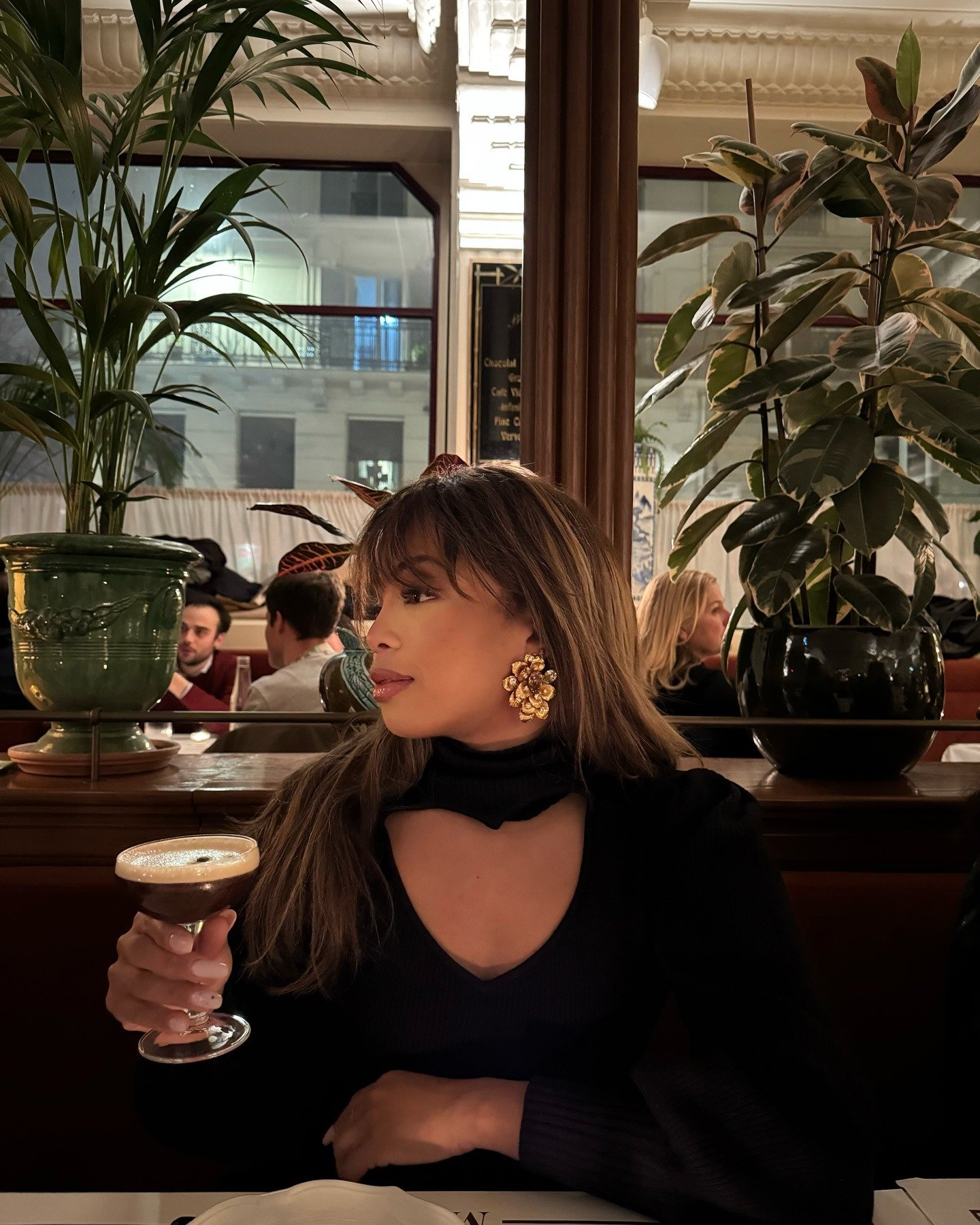 Mulher posa segurando taça, usando maxi brinco dourado floral, blusa preta com recorte e ambiente sofisticado, acessórios