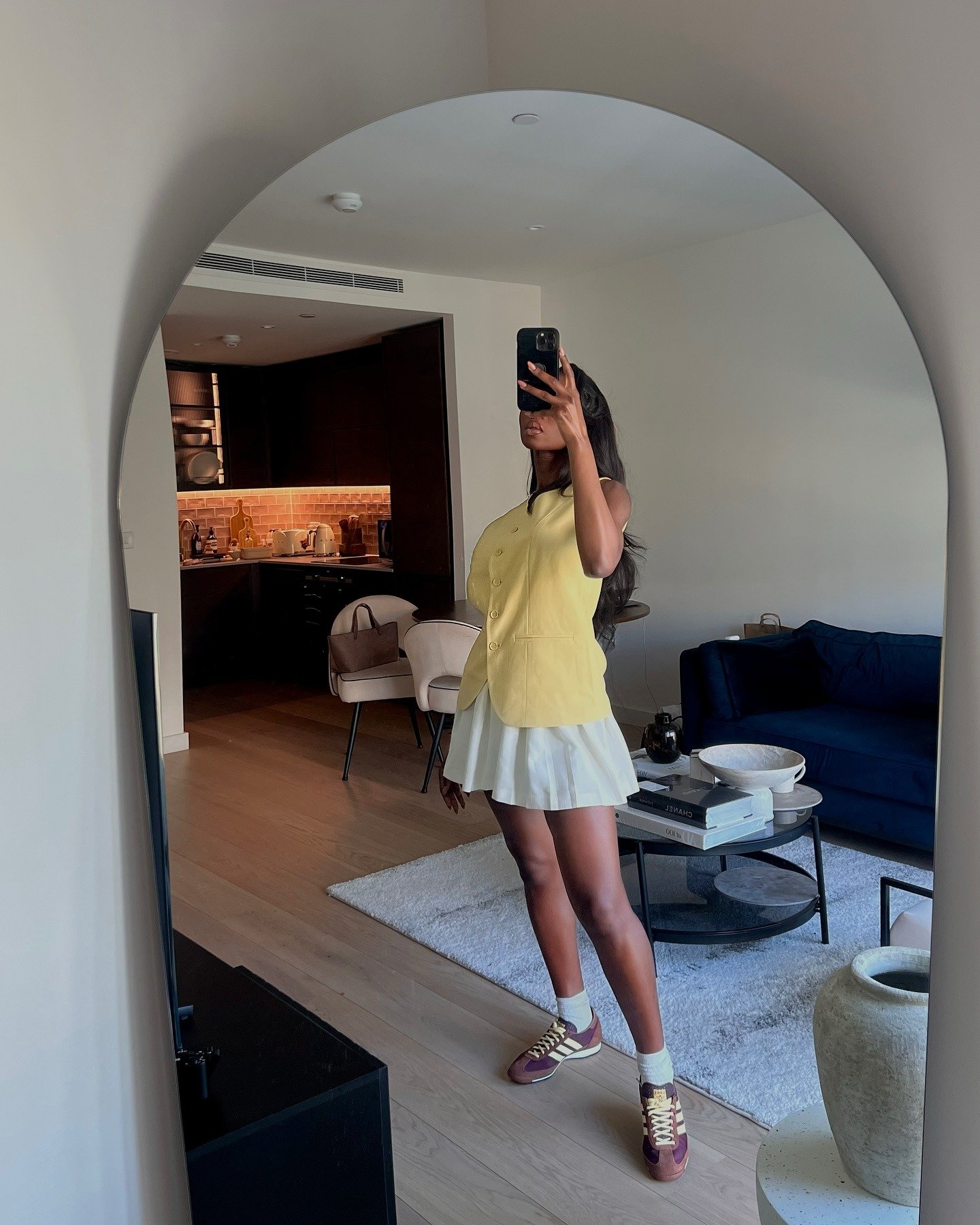 Modelo posa em frente a espelho com tênis adidas feminino vinho e bege, saia plissada branca e colete amarelo.