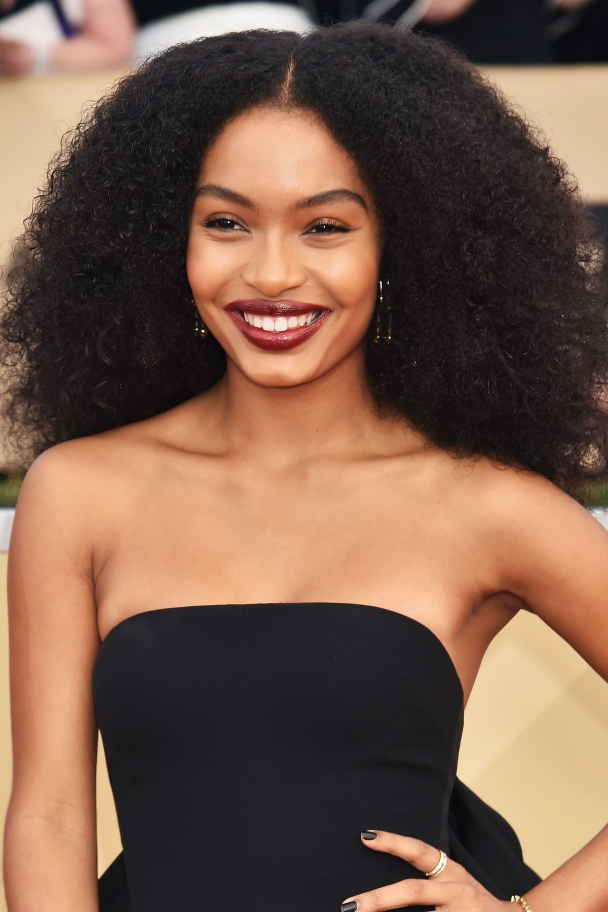 Modelo sorri confiante com penteados afro volumosos e naturais, usando vestido tomara que caia preto e batom vinho intenso.