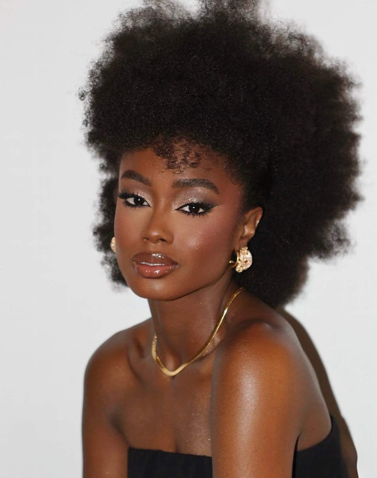 Modelo posa com penteados afro volumosos e naturais, combinando acessórios dourados e maquiagem iluminada em fundo neutro.