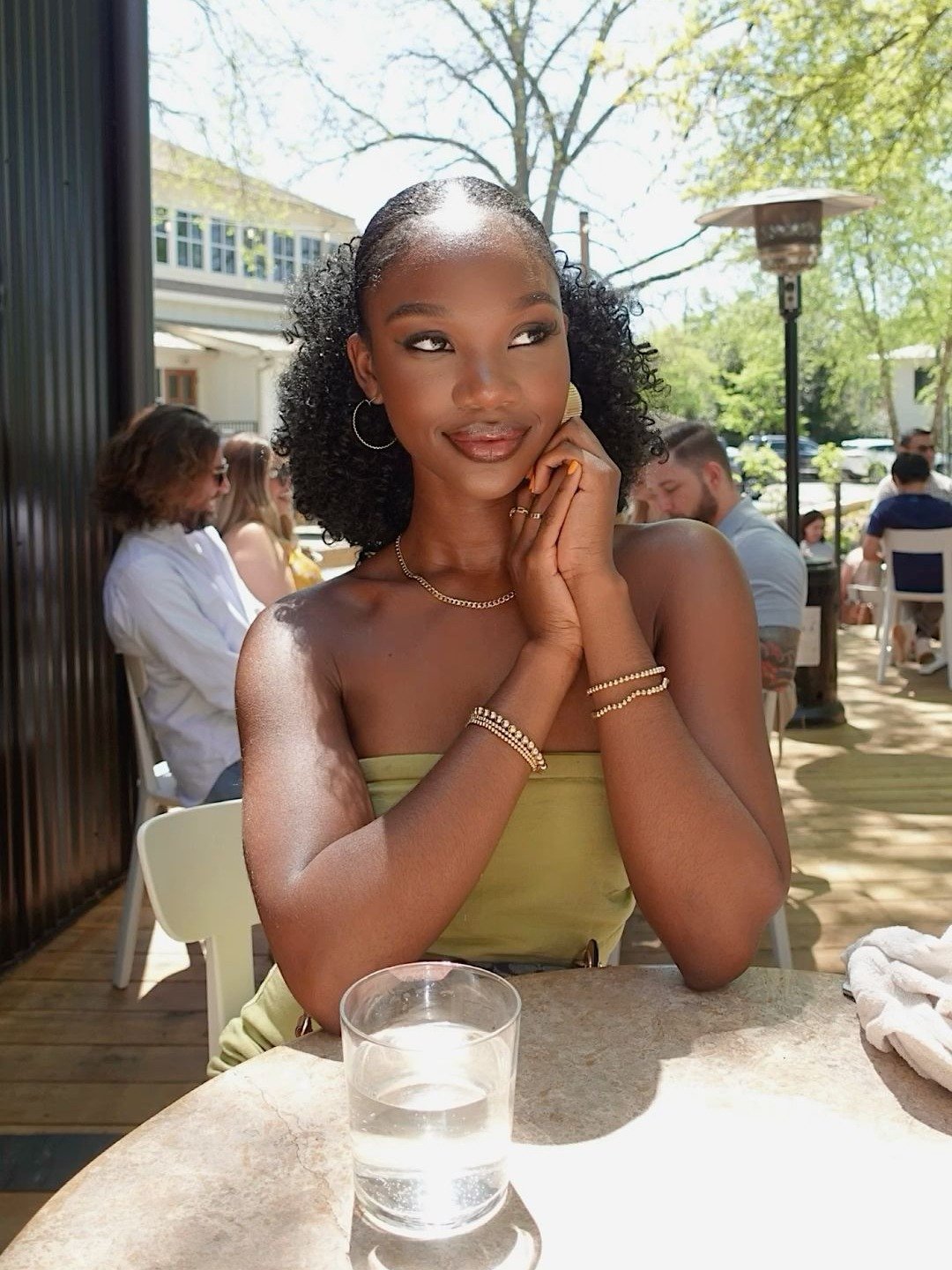 Modelo com penteado afro volumoso preso atrás, vestido tomara que caia verde oliva e acessórios dourados.