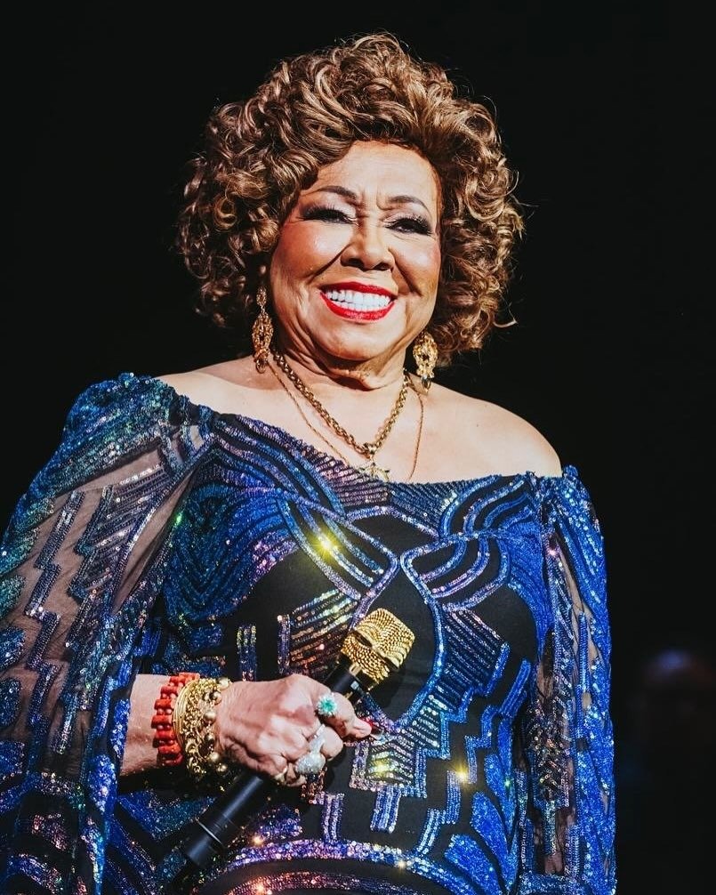 Alcione sorri radiante segurando microfone dourado, veste vestido azul com bordados brilhantes e mangas transparentes.