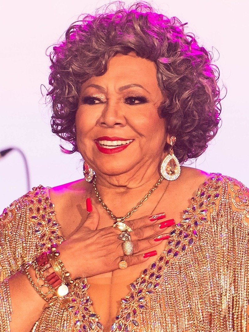 Alcione sorri com emoção, usando vestido dourado bordado, joias brilhantes, unhas vermelhas e cabelo cacheado.
