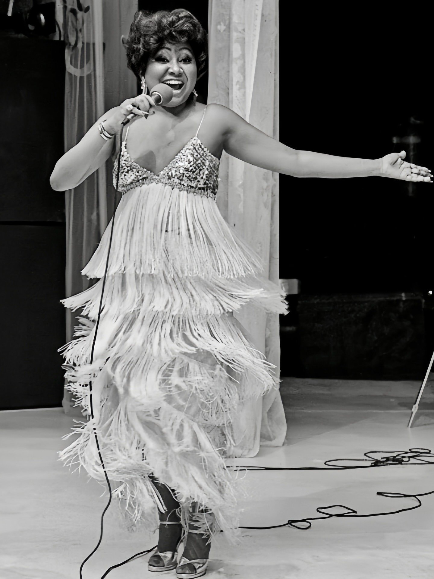 Alcione dança animada com vestido de franjas e brilho, modelagem fluida e alças finas, em clima vibrante e expressivo.