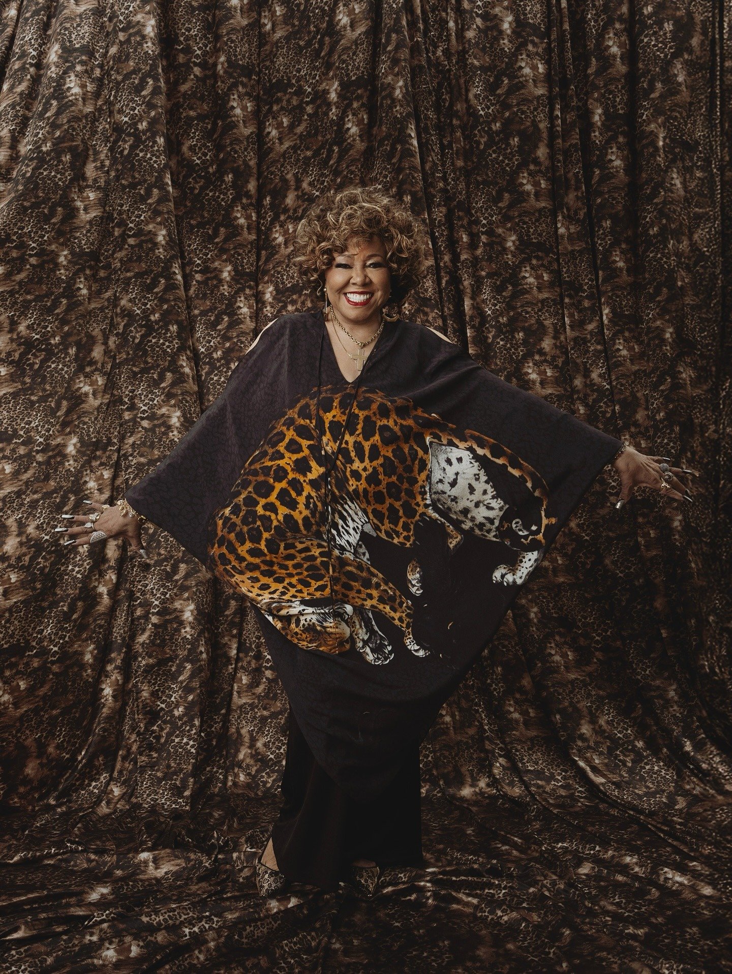 Alcione sorri radiante, vestindo túnica preta com estampa de onça vibrante e fundo animal print marrom.