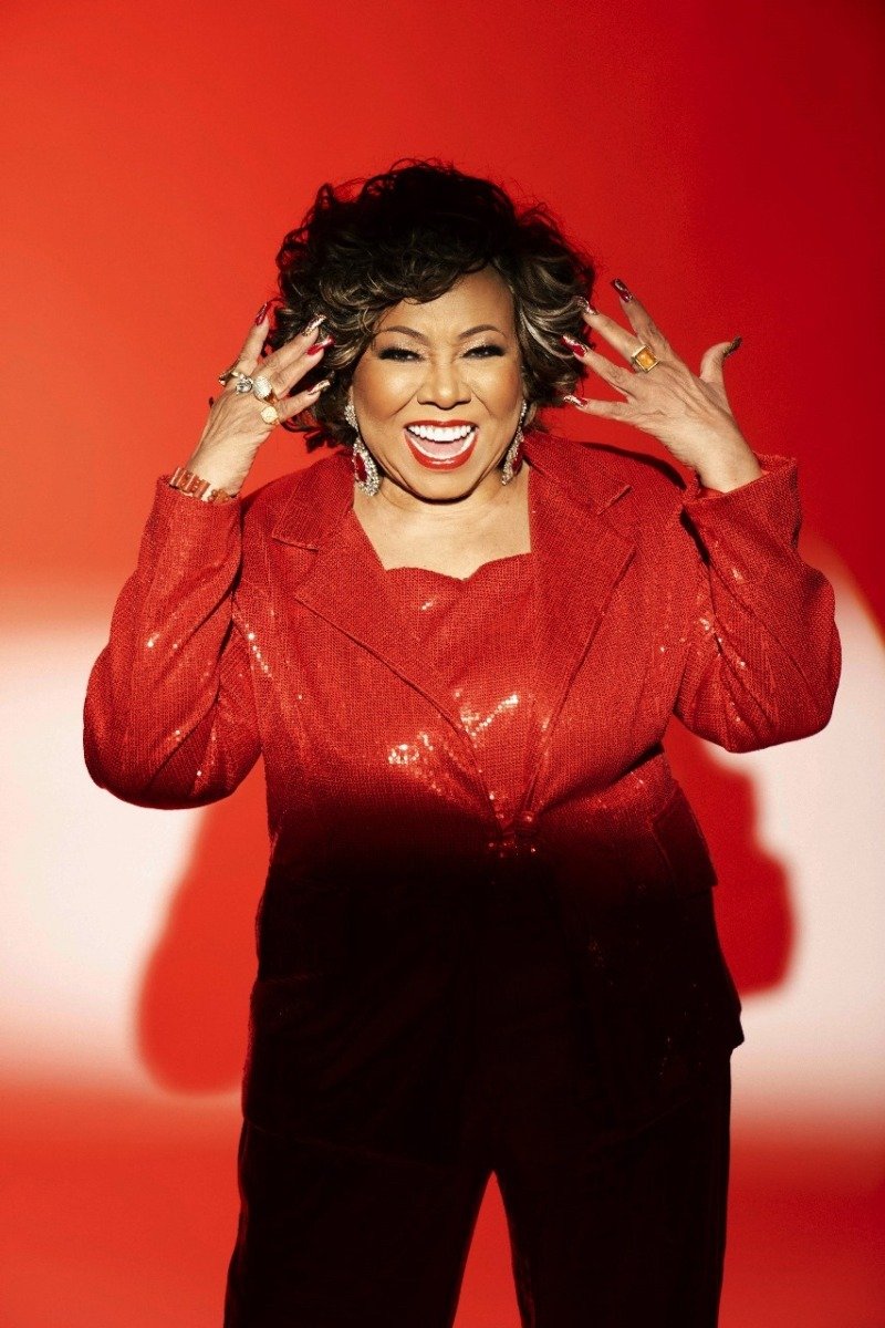 Alcione sorri radiante e posa com energia, vestindo conjunto vermelho de paetês com blazer estruturado e acessórios dourados.