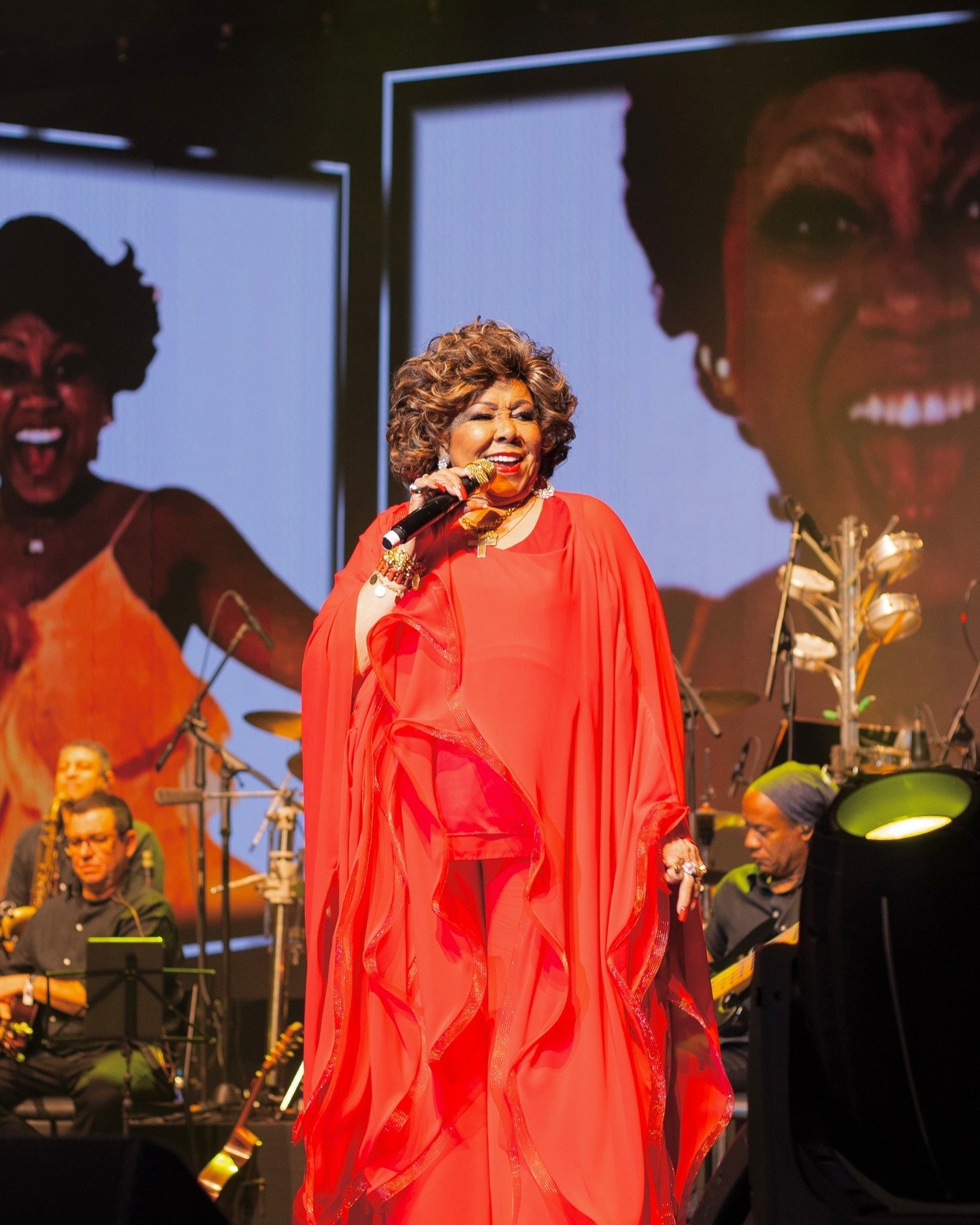 Alcione canta no palco com vestido fluido vermelho vibrante em tecido leve, transmitindo energia e alegria contagiante.