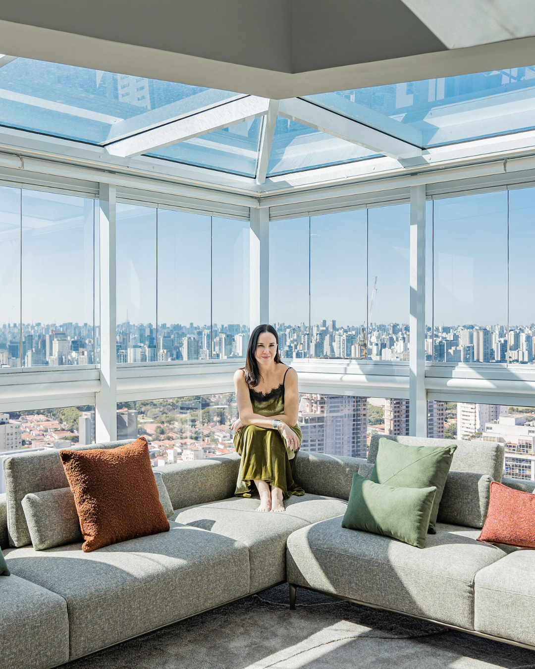 Ambiente de decoração 2026 com sofá modular cinza, almofadas verdes e terrosas, iluminação natural e modelo sentada relaxada