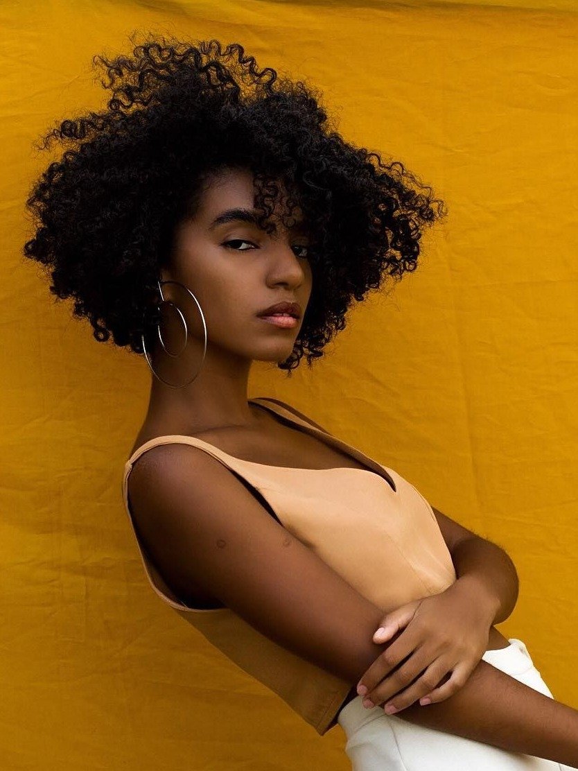 Corte de cabelo 2026 com volume natural e cachos definidos, modelo posa confiante em blusa bege e fundo amarelo vibrante.