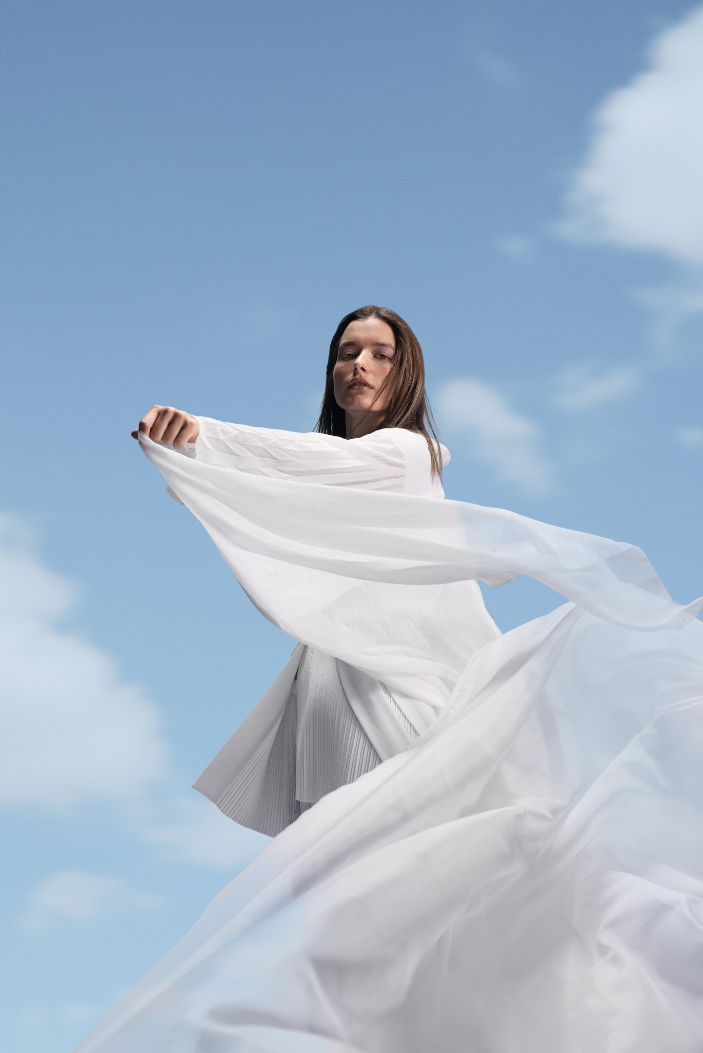 Modelo posa com elegância usando peças brancas fluidas e plissadas em tecido leve, sob céu azul claro, transmitindo leveza e