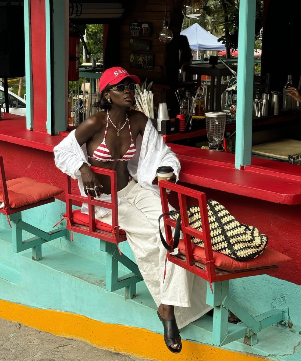 Modelo senta com postura confiante em banco vermelho, usando biquíni listrado, calça branca e bolsa para praia trançada.