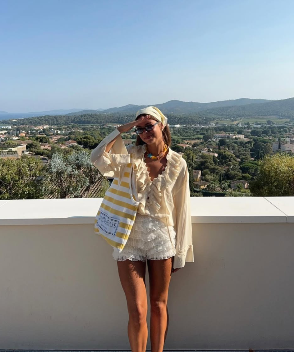 Modelo posa com blusa creme de babados, shorts rendados brancos e bolsa para praia listrada amarela em cenário ensolarado.